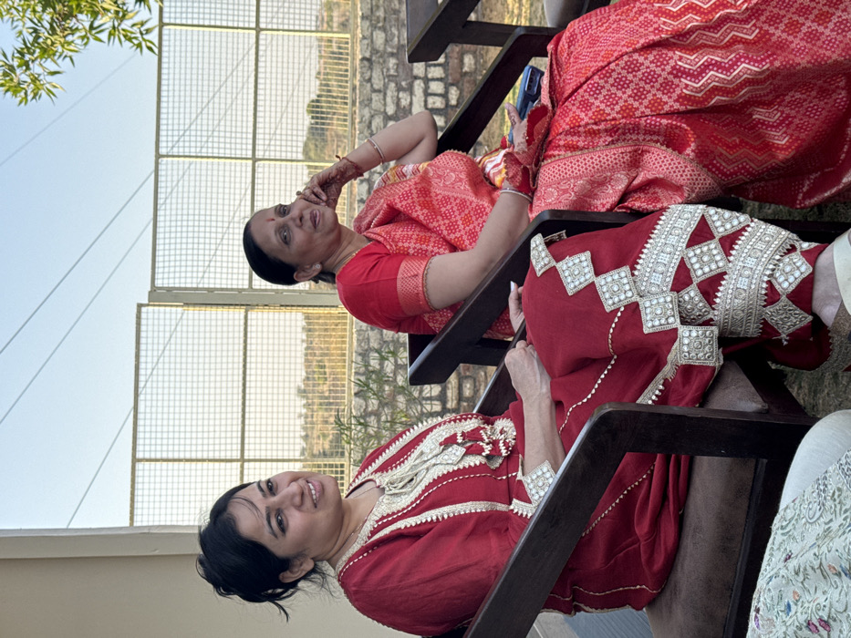 Two women are seated outdoors in Malasar, India, under a clear, pale blue sky. The woman on the left, with dark hair tied back, wears a long-sleeved, deep red garment featuring intricate gold and white embroidery and tasselled ornamentation around the neckline and on the lower legs. She is smiling and looking towards the right. The woman on the right, with dark hair pulled back, wears a patterned red saree with gold accents. She is also smiling and looking towards the right, with her right hand, adorned with mehndi, resting near her face. Both women occupy dark-brown wooden chairs with upholstered seats. A blue mobile phone is partially visible on the lap of the woman on the right. In the background, a light beige building wall is visible to the left. A metal grid fence separates the immediate seating area from a distant, arid landscape characterized by sparse vegetation and low hills. A low stone wall is visible beneath the fence to the right. Overhead, utility lines traverse the sky, and green foliage is present on the far right.