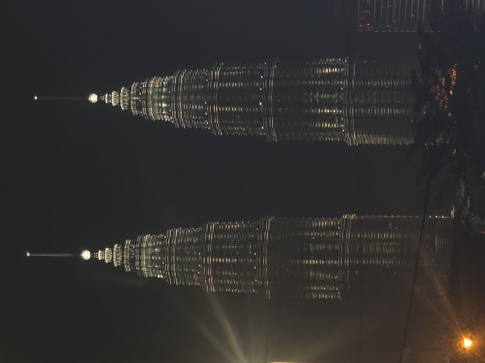 The image shows the Petronas Twin Towers reflected on a dark surface, likely water or a reflective window, at night. The towers are illuminated with lights, creating a mirror image. A building can be seen in the top right corner. The location is Kuala Lumpur, Malaysia, where the Petronas Towers are located.