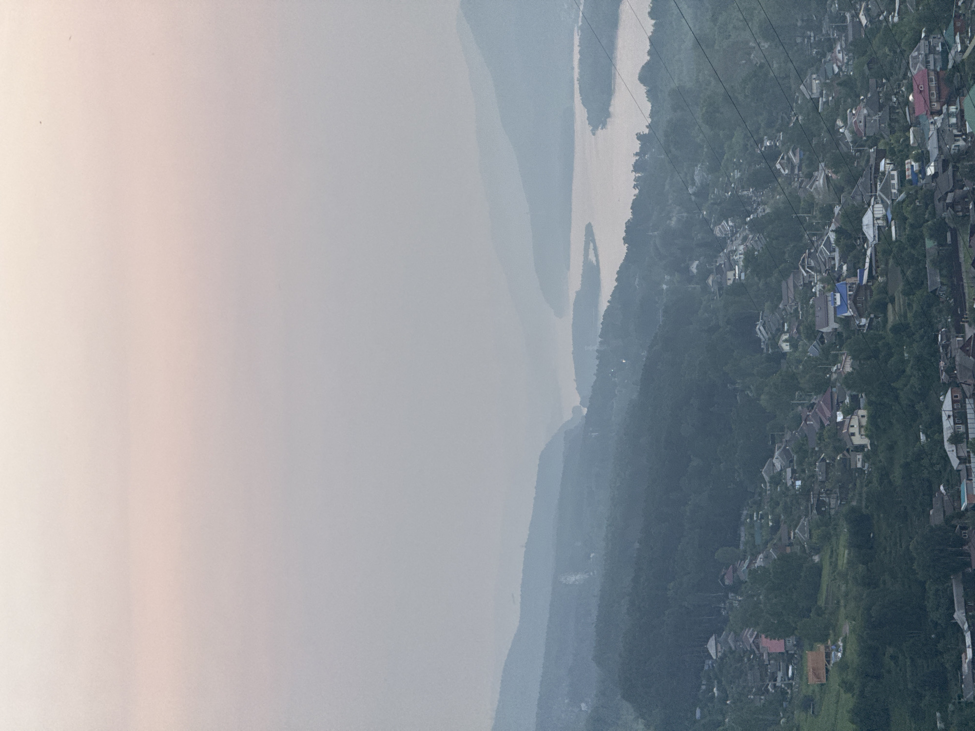 This media file captures an aerial or elevated view of a sprawling settlement nestled on a hillside overlooking a winding river. The scene is characterized by a soft, hazy atmosphere, suggesting it could be early morning or a day with mist or fog. The river, appearing lighter in color, snakes through the landscape, flanked by the dense, dark green foliage of a forest that covers the rolling hills.
The settlement itself is a dense collection of houses with varied roof colors, including red, gray, and blue, interspersed with trees. Some structures appear to be homes, while others might be commercial buildings. The architecture suggests a somewhat dense, perhaps developing urban or semi-urban area, built into the natural terrain. Power lines are visibly strung across the frame, indicating the presence of infrastructure.
The overall impression is one of a community integrated with its natural environment, with the river and hills providing a dramatic backdrop. The hazy conditions create a sense of depth and distance, with layers of hills fading into a soft, diffused light on the left side of the image. The time of day is ambiguous due to the haze, but the subdued lighting leans towards dawn or dusk. There is no visible text in the image. The setting appears to be a remote or mountainous region, given the natural landscape and the settlement's placement.
