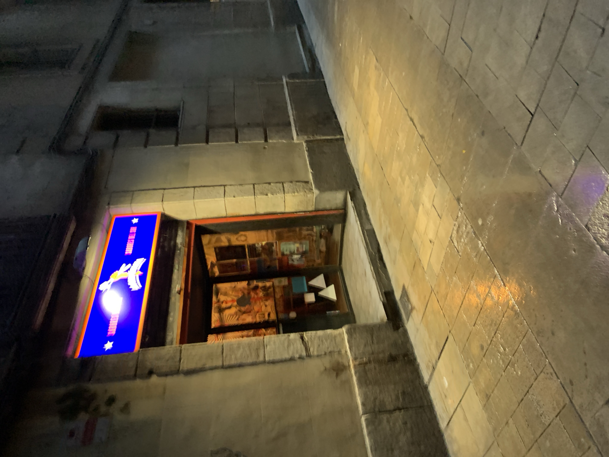 The image depicts the exterior of a shop located in Barcelona, Spain. The shop has a blue illuminated sign with a stylized graphic of a bird or winged figure above the words "THE" and "BARCELONA". The storefront displays a variety of objects, including what appear to be sculptures or decorative items on tables. The architecture is characterized by stone masonry and a narrow street. The street's surface is reflective, possibly due to recent rain, and shows a pattern of rectangular paving stones. The scene is lit by streetlights.