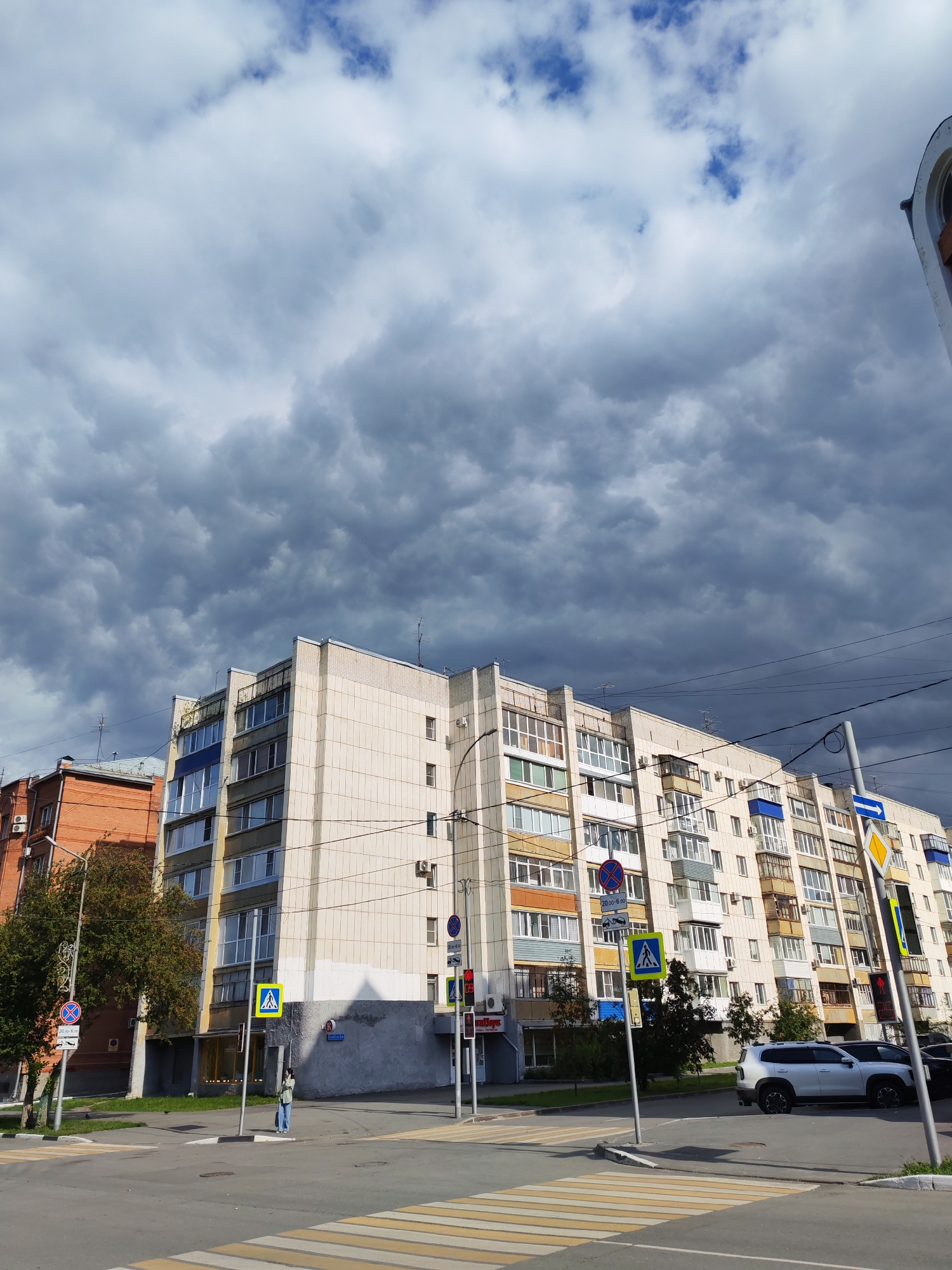 A wide-angle shot captures a street scene under a cloudy sky. In the foreground, a pedestrian crossing with yellow and white stripes is visible. A light-colored SUV and a black car are parked on the right side of the street. A multi-story residential building, with a cream-colored facade and blue and white window accents, dominates the background. To the left of this building is a shorter, red-brick building. Street signs, including pedestrian crossing and directional signs, are present throughout the scene. A lone pedestrian stands on the sidewalk near a sign. The sky is filled with dramatic, dark gray clouds, suggesting an impending storm.