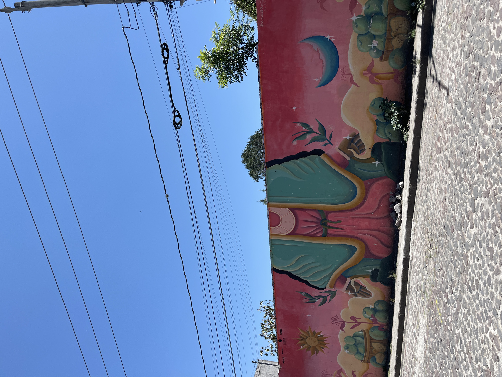 A vibrant outdoor scene in San Andrés Cholula, Mexico, features a colorful mural, utility lines, and a cobblestone path under a bright blue sky. The central focus is a vertical wall adorned with a mural, predominantly in pinkish-red.
The mural depicts a large, stylized figure in flowing teal and pink garments, reminiscent of the Virgin Mary, though only the upper body is partially visible. Above, a light blue crescent moon is surrounded by small stars, while a yellow sun with rays is painted towards the lower left. Baskets overflowing with green fruits, possibly apples, are interspersed with green foliage around the central figure. Faint text visible on the mural includes "@KAPINGBERN", "Comex", "AURORA", "MEXICO", and "2023".
To the right of the mural, a textured cobblestone path or sidewalk, composed of dark, rounded stones, runs alongside the wall. Above the mural, against a clear, cloudless blue sky, several black utility lines stretch horizontally, some connected to visible pole sections and insulators. Green tree foliage can be seen peeking over the wall and beneath the utility lines. The scene is illuminated by bright daylight, indicating a sunny day.