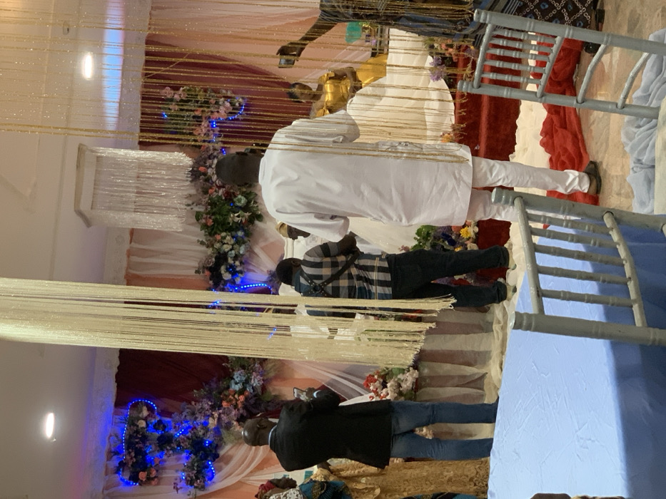 A wedding scene includes floral decorations and beaded curtains. Several people are standing around. One man wears a white outfit with matching pants. Another man is in a plaid shirt with jeans, and a third man wears a black jacket. Chairs with a blue tablecloth can also be seen. The photo appears to be taken indoors, possibly at a reception hall.