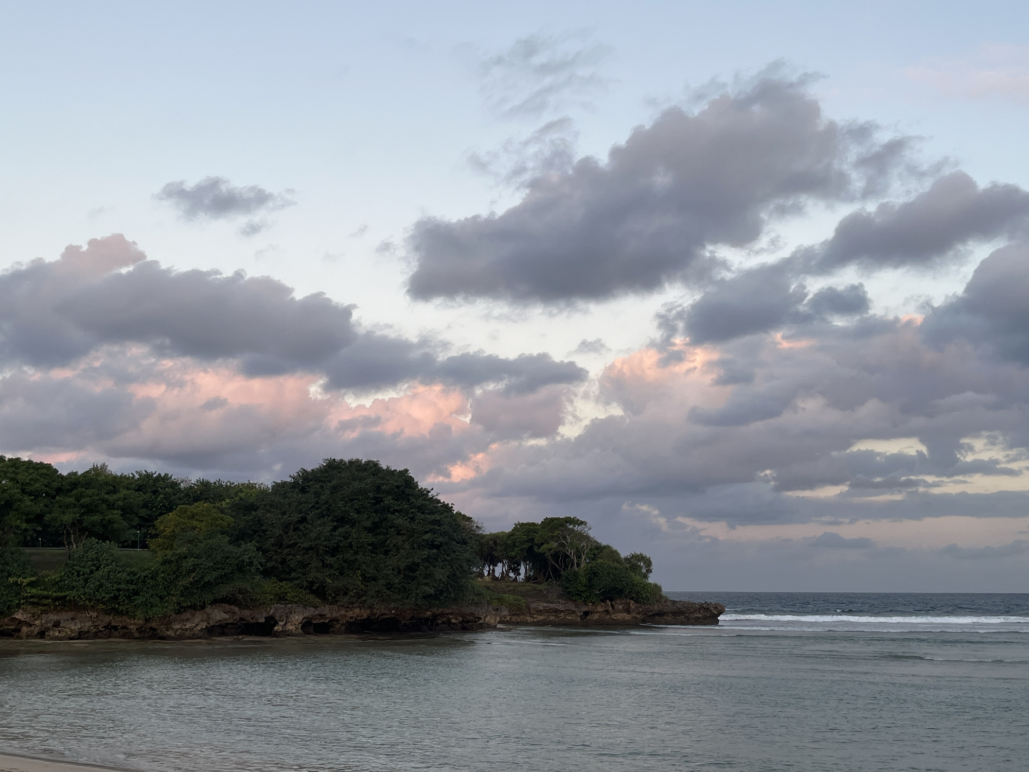 The image depicts a tranquil coastal scene in Kuta Selatan, Indonesia, during what appears to be dusk or dawn. The sky is a gradient of soft blues and grays, punctuated by large, puffy clouds tinged with pink and lavender from the setting or rising sun. Below the sky, a lush, green coastline is visible, characterized by dense foliage and rocky outcrops. The vegetation is thick and varied, suggesting a tropical environment. The ocean in the foreground is calm, with gentle waves rolling towards a sandy shore, which is partially visible at the bottom of the frame. The water transitions from a clear, shallow area near the shore to a deeper blue further out. No people or man-made structures are prominently featured, emphasizing the natural beauty of the location. The overall atmosphere is serene and picturesque, characteristic of a quiet beach in Kuta Selatan.
