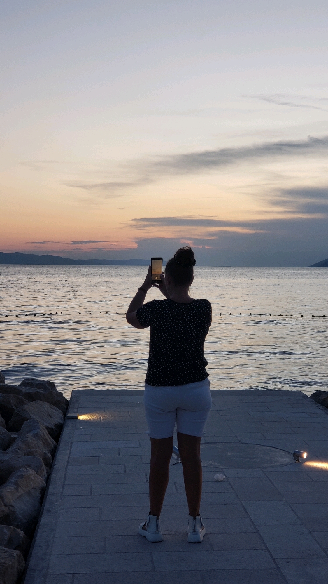 A person stands on a paved walkway at dusk, facing a body of water and capturing a photograph with a smartphone. The person is seen from behind, wearing a dark, polka-dotted short-sleeved shirt and white shorts. They have their hair pulled up in a bun. The walkway appears to be made of gray pavers and is lined with large rocks on the left. In the background, the sea stretches to the horizon, with a faint outline of mountains visible in the distance. The sky displays the soft colors of sunset, with hues of pink, orange, and gray clouds. The scene suggests a tranquil evening by the coast, likely in a town like Općina Tučepi, Croatia, known for its coastal beauty.