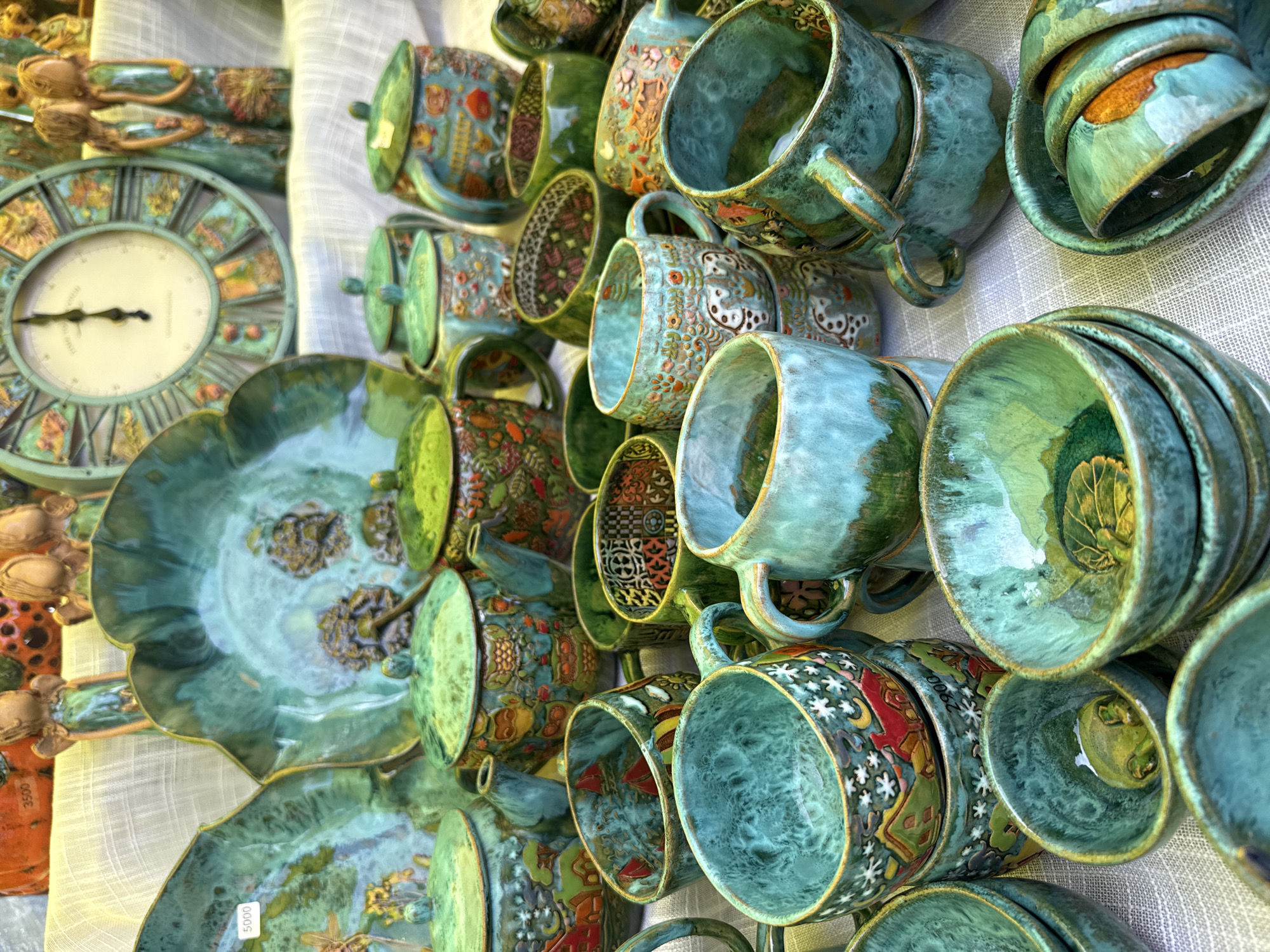 In Старые Быги, Russia, a close-up, high-angle shot captures a vibrant display of handcrafted ceramic pottery arranged on a white linen tablecloth. The collection features an assortment of teapots, cups, and bowls, all adorned with intricate designs and finished in rich shades of turquoise, emerald green, and earthy brown. Some pieces are decorated with motifs of flowers, animals, and whimsical scenes, while others boast a more abstract, glazed finish. The lighting is bright and diffused, highlighting the textures and colors of the ceramics. In the background, out-of-focus elements suggest a market stall or a decorative display, with a large clock face visible above the pottery. No people are visible in the frame.