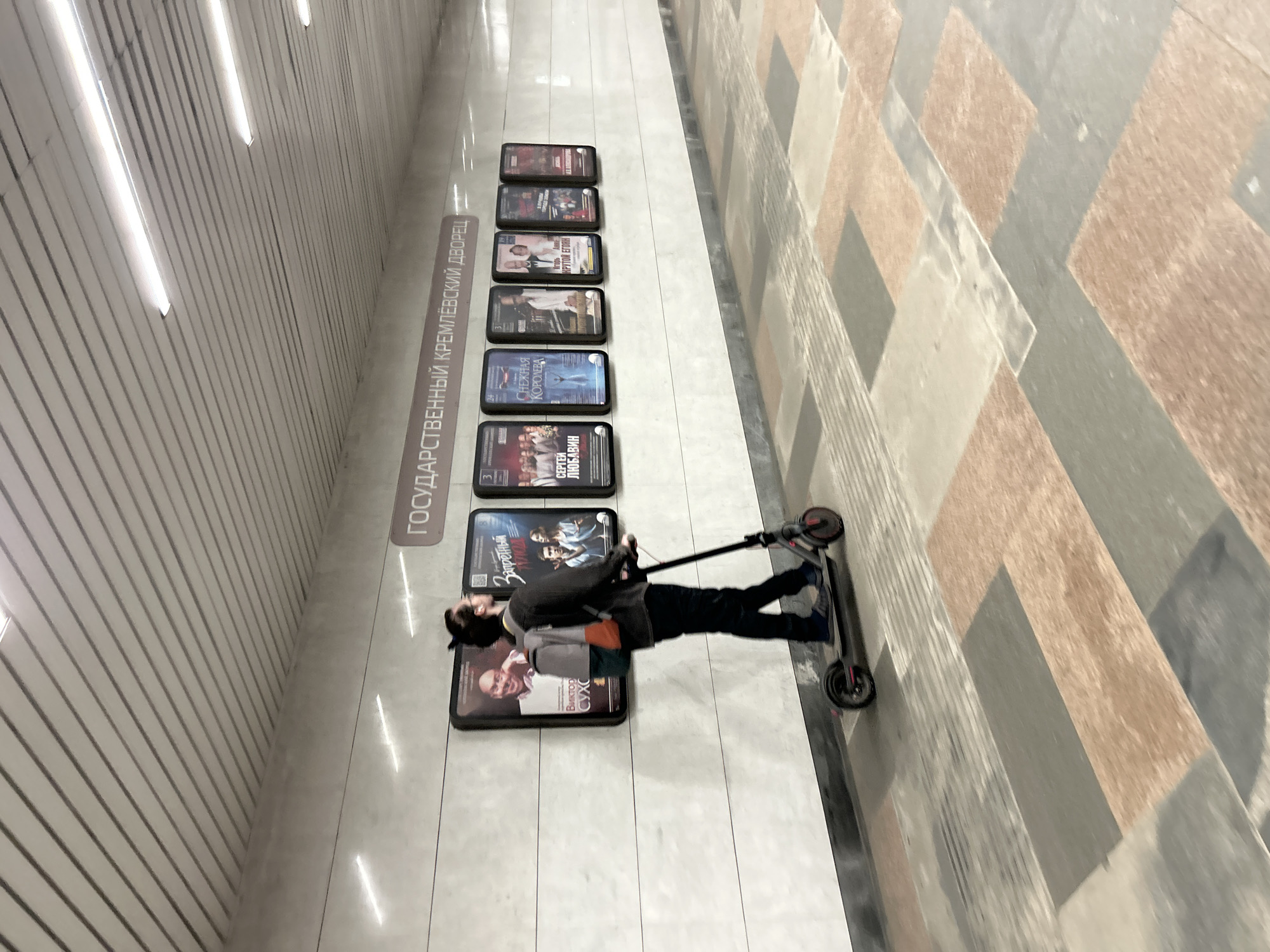 A full shot captures a person riding an electric scooter down a subway station hallway. The person, viewed from the right, has dark hair pulled back in a bun and wears glasses, a dark long-sleeved shirt, dark pants, and grey slip-on shoes. They also carry a backpack with grey and orange accents. The scooter is dark grey with red accents on the wheels. The hallway walls are lined with framed posters featuring theatrical or cultural events. Above the posters, a sign reads "ГОСУДАРСТВЕННЫЙ КРЕМЛЁВСКИЙ ДВОРЕЦ" (State Kremlin Palace). The floor is tiled in a pattern of rectangular blocks in grey, beige, and reddish-brown hues. The ceiling is white with parallel, recessed lights.