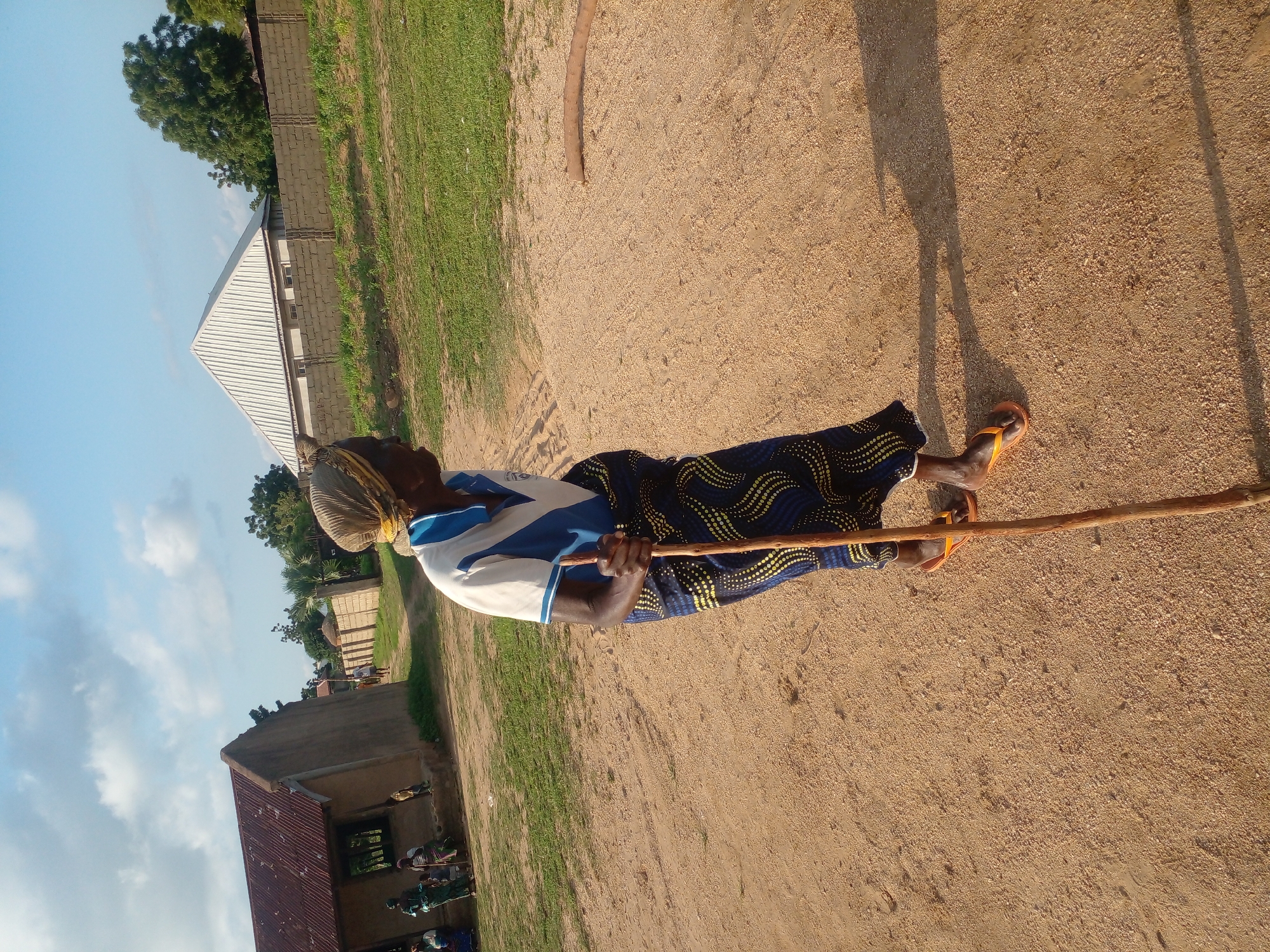 An elderly woman, wearing a headscarf, a blue and white polo shirt, and a patterned skirt, is standing on a dirt path and holding a wooden walking stick. She is looking to her right. In the background, there is a building with a red corrugated iron roof, other people, a fence, and trees. The sky is partly cloudy and blue. The scene appears to be in an outdoor, rural or semi-rural setting, possibly in the city of Langtang, Nigeria.