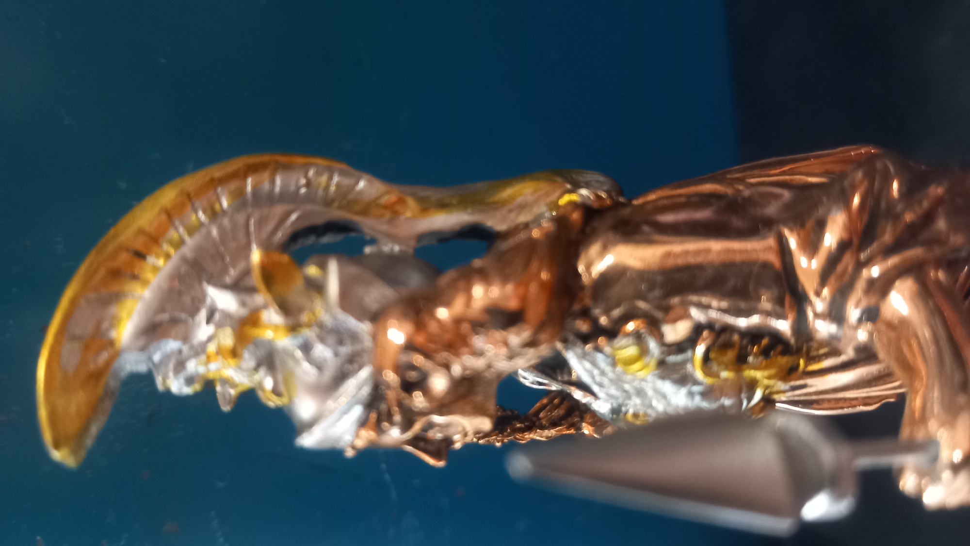 The image shows a close-up, side-profile view of a metallic figurine, possibly a statue or a collectible. The figurine is adorned with intricate details, including a helmet with a prominent crest and what appears to be a beard or braided hair. The metallic finish suggests it might be made of bronze or a similar material, with highlights reflecting light. The background is a solid, deep blue. No specific location in the city of Kos, Greece, can be identified from this image.