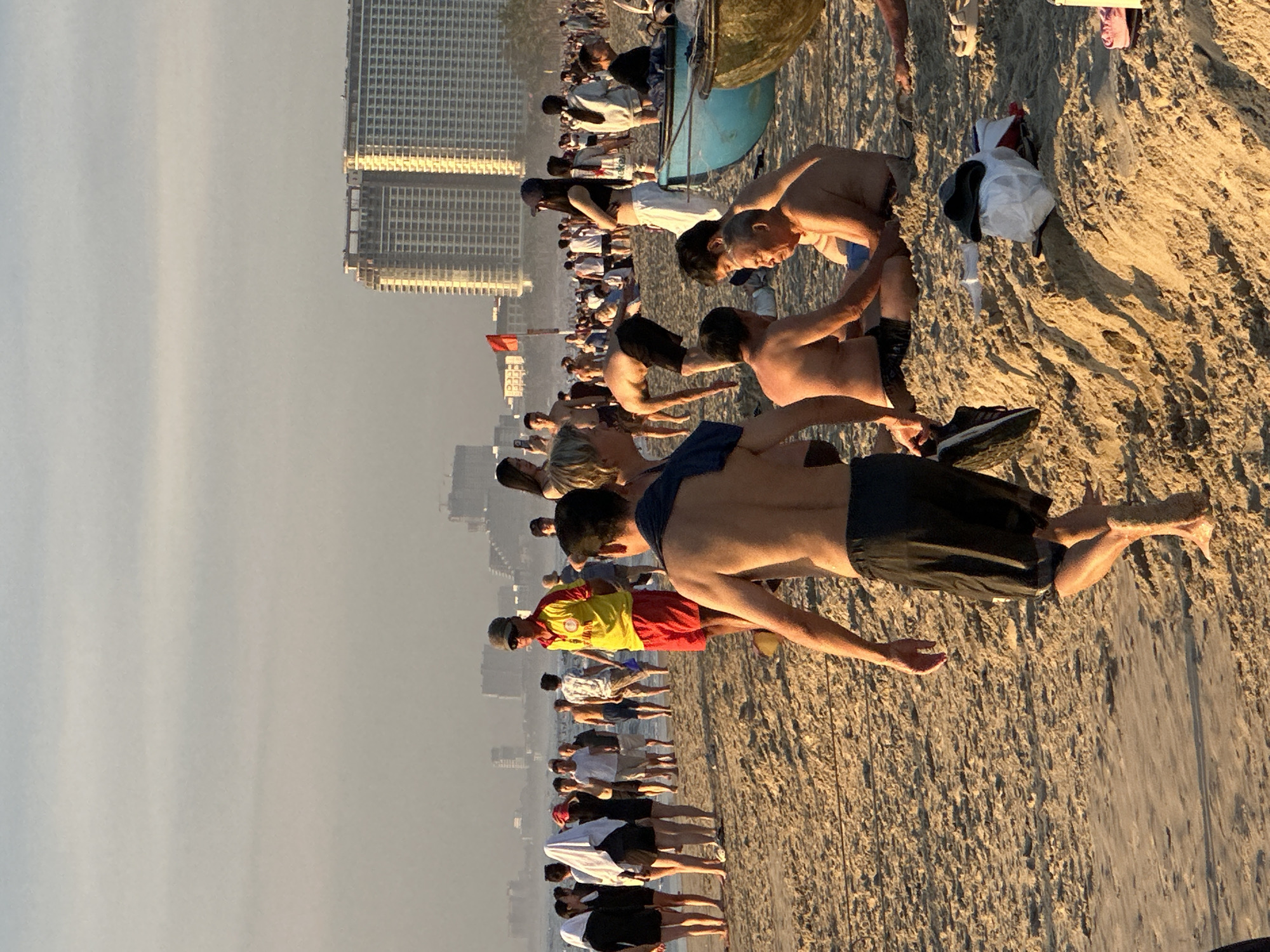 This image shows a large group of people on a sandy beach, likely during an outdoor exercise or training session. The subjects are diverse in age and gender, mostly dressed in athletic wear or casual beach attire. They are arranged in rows, facing the sea, and appear to be engaged in synchronized movements, possibly stretching or a form of martial arts or fitness routine.
In the background, tall buildings are visible, suggesting an urban coastal setting. The sky is hazy and appears to be either early morning or late afternoon, as indicated by the warm, low-angled sunlight casting long shadows on the sand. The weather seems to be clear, albeit with some atmospheric haze.
Notable details include a red flag planted in the sand in the middle distance, which might signify a marker or a point of interest for the group. There's also a blue boat-like object and some scattered items on the sand, including bags and shoes, suggesting people have come prepared for an activity. The general atmosphere seems energetic and focused, with individuals concentrating on their movements. The location is identified as Đà Nẵng, Vietnam, which aligns with the presence of high-rise buildings along a beach. No discernible text is visible in the image.