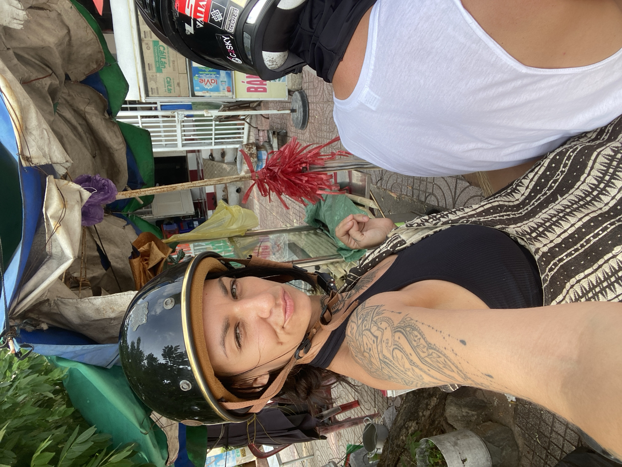 A woman with dark hair and tattoos on her arm and shoulder is riding on the back of a motorcycle, looking over her shoulder at the camera. She is wearing a black helmet with a gold trim and a black tank top. Her face is partially obscured by the helmet. The motorcycle appears to be stopped on a street with a makeshift stall or shelter behind her, covered with various tarps and fabrics. In the background, there are shelves with products, possibly from a store, and a white gate. Another person, wearing a white t-shirt and a black helmet, is sitting in front of her. The environment suggests a tropical urban setting, likely in Mũi Né, Vietnam, given the style of the shelter and the presence of what appears to be local storefronts. The overall lighting indicates it is daytime.