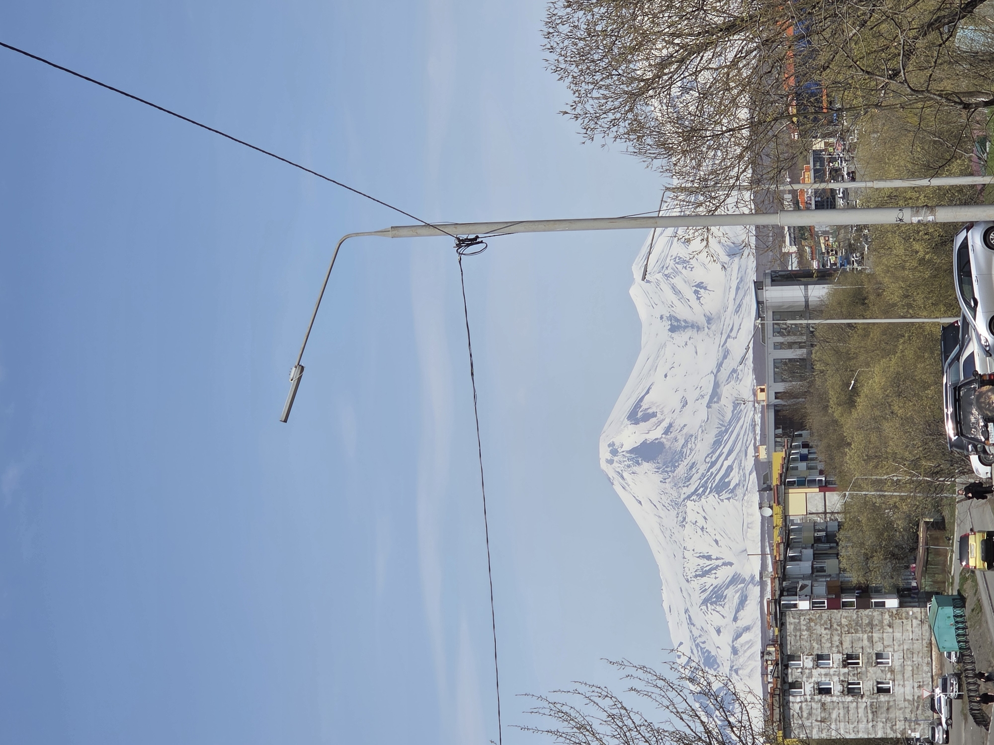 The image shows an urban street scene with a prominent snow-covered mountain in the background. A street light pole is visible in the foreground, with power lines running across the sky. Buildings of various heights and colors line the street, interspersed with trees displaying early spring foliage. Cars are parked along the side of the road and some pedestrians are walking in the distance.