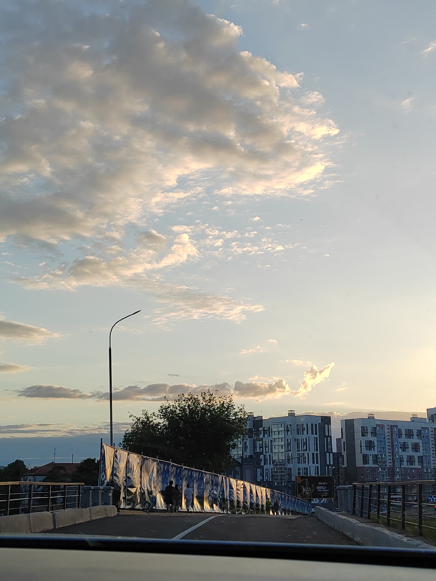 The image depicts a view from a vehicle on a bridge, looking forward. The sky is filled with a mix of white and grey clouds, illuminated by what appears to be sunset or sunrise light. In the distance, modern apartment buildings with numerous windows are visible. Along the side of the bridge, there is a temporary barrier covered with large, patterned fabric banners, possibly for construction or an event. A lone street lamp stands on the left side of the bridge. A few people are walking on the bridge, partially obscured by the banners. The environment suggests an urban setting in Brest, Belarus, with elements of contemporary architecture and infrastructure.