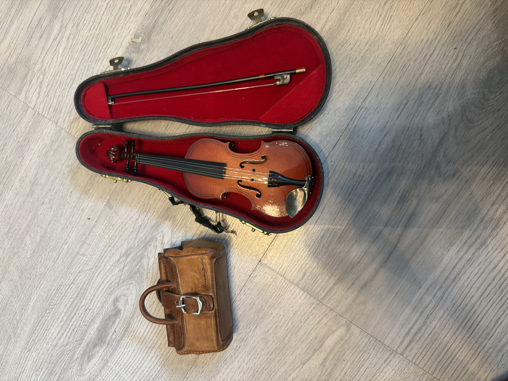 An overhead shot shows a miniature brown leather case with a buckle and handle next to an open black violin case lined with red velvet. A miniature violin with a bow rests inside the open case. The objects are positioned on a light gray laminate wood floor with a wood grain pattern.