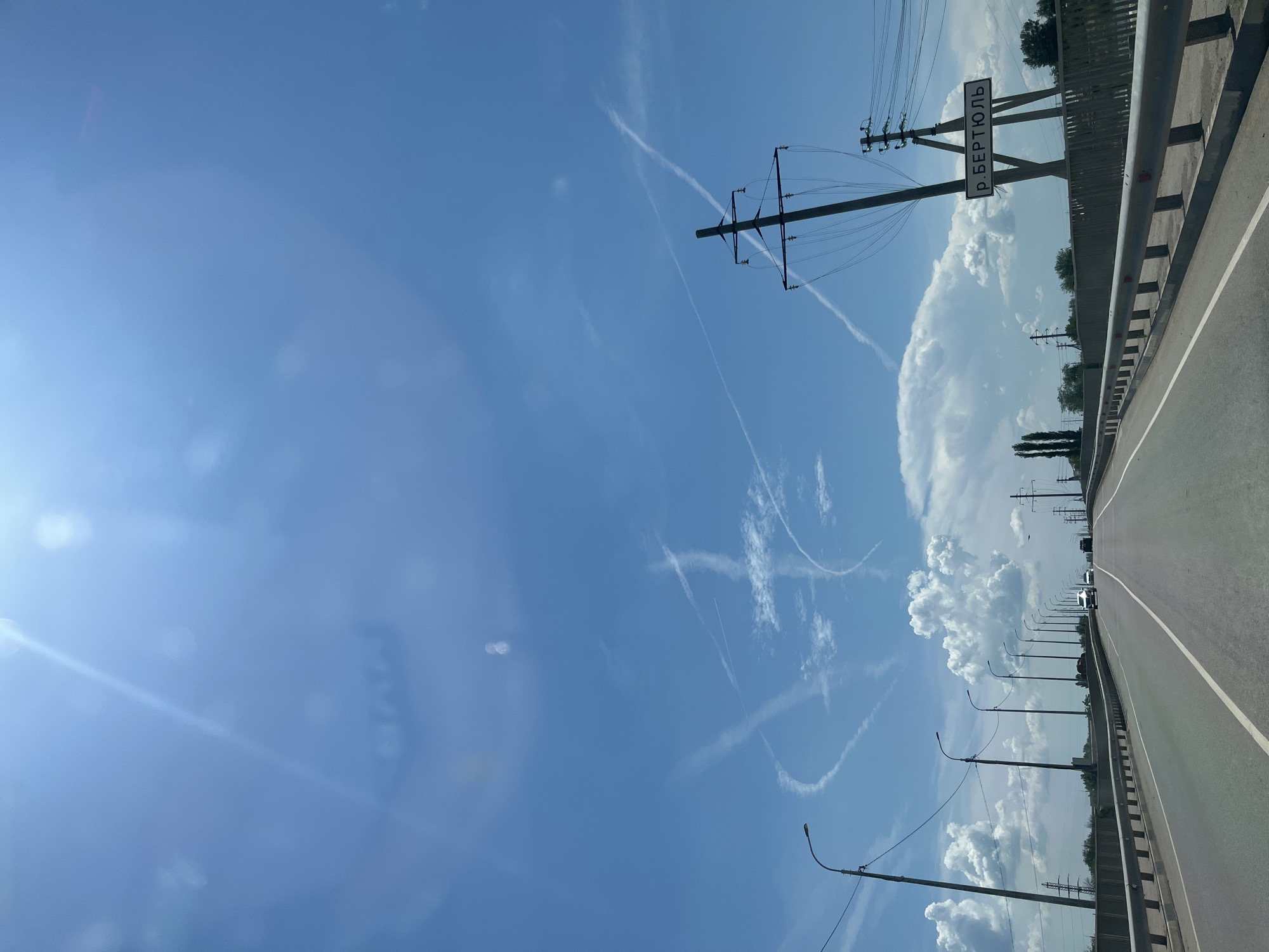 The image shows a road crossing a bridge, likely in Krasnyye Barrikady, Russia. The sky is blue with scattered clouds and contrails from airplanes. There are several streetlights and power lines visible along the road. On the right side of the road, a sign reads "р. БЕРТЮЛЬ," which is likely a geographical marker for the Bertiul River. A few vehicles are on the road, including cars and trucks, suggesting moderate traffic. The overall scene is typical of a rural or suburban highway environment.