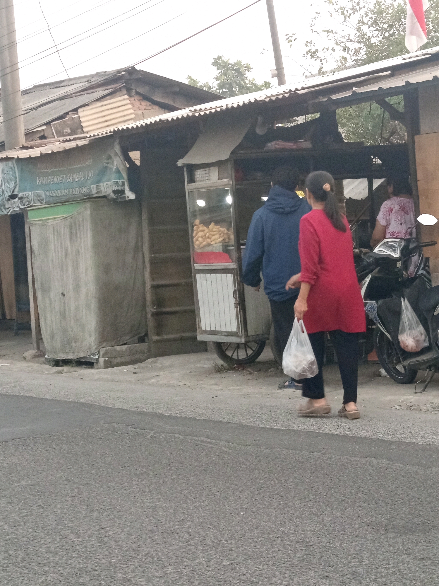 A street-level, outdoor, medium-shot captures a scene in Karawang, Indonesia, where an outdoor food stall operates on a slightly overcast day. In the foreground, a paved road takes up the bottom half of the frame. Behind the road, a grey concrete curb separates the street from a dirt and gravel area. A woman in a red long-sleeved shirt and black pants, carrying a clear plastic bag filled with items, stands with her back to the viewer, facing a food cart. To her left, a man in a blue hooded jacket, also with his back to the viewer, stands at the food cart. The food cart, made of metal and glass, displays fried items in its upper section and has shelves underneath. To the right of the food cart, a parked motorcycle is visible, with another person in a patterned shirt standing near it. Behind the food stall, the structures appear to be made of corrugated metal and wood, with a weathered, rustic aesthetic. A partially covered, grey, rectangular structure is situated to the left of the food cart, possibly a storage area. The background reveals foliage and the cloudy sky. The overall impression is of a typical, bustling roadside eatery in an Indonesian city.