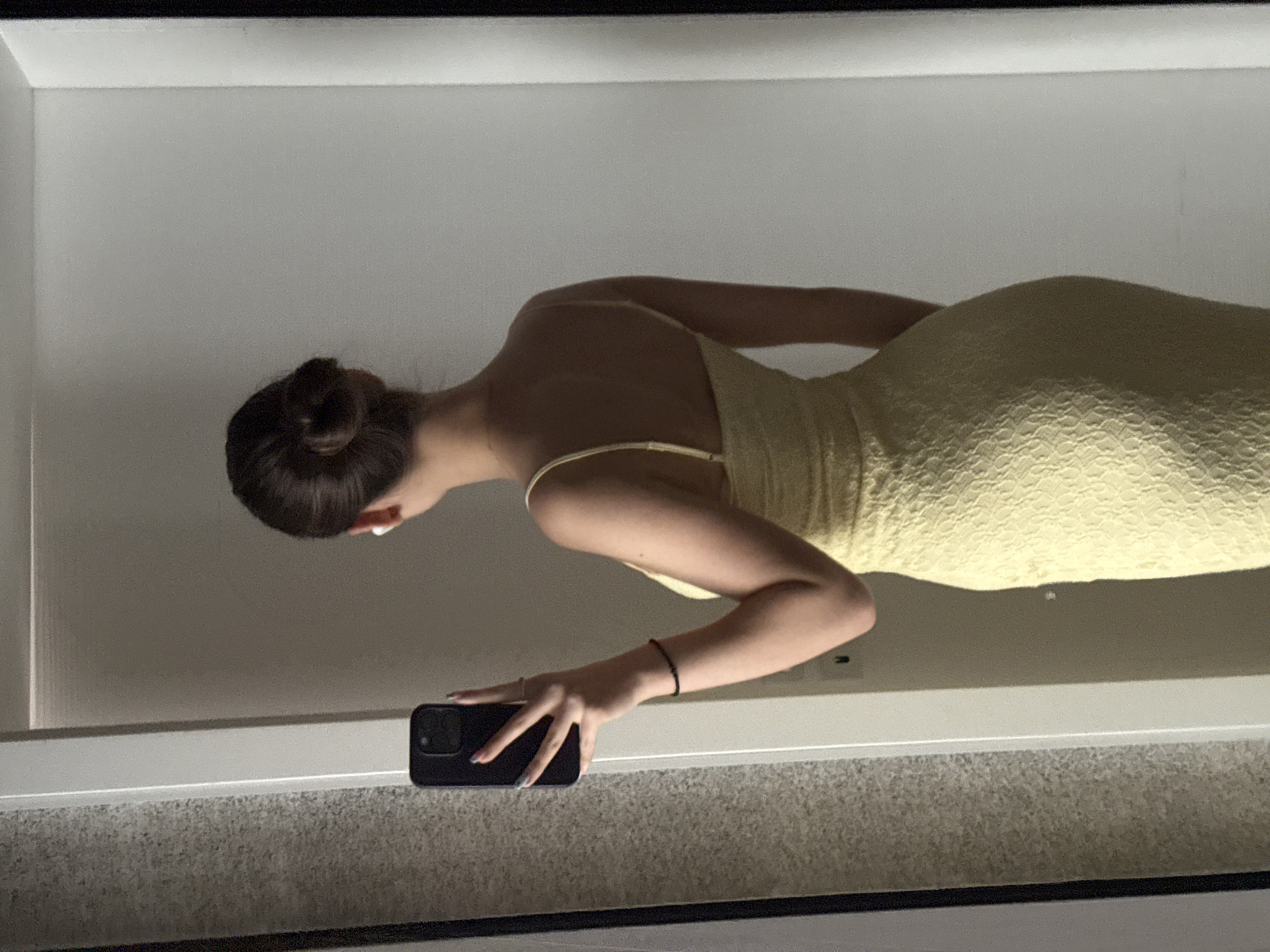 The image is a mirror selfie taken indoors, featuring a person in a light yellow, textured, spaghetti-strap dress. The person has dark hair pulled back into a bun and is captured from the back and side, leaning slightly forward and looking down. Their face is not visible.
In the reflection, the person's right arm is bent, and they are holding a dark-colored smartphone, likely an iPhone, with their right hand to take the photo. The fingernails of the hand holding the phone appear to have light-colored polish. A thin, dark bracelet is visible on their right wrist.
The background visible in the mirror reflection consists of a plain, light-colored wall or surface, possibly part of a wardrobe or room interior. A prominent white horizontal strip, possibly a shelf edge or door frame, runs across the lower part of the reflection. Below this strip, a grey, textured surface, likely a carpet or rug, is visible. The overall lighting suggests an indoor setting, possibly with artificial light or diffused natural light, as there are no harsh shadows. There is no visible text in the image. The location context provided for this indoor scene is Sofia, Bulgaria.