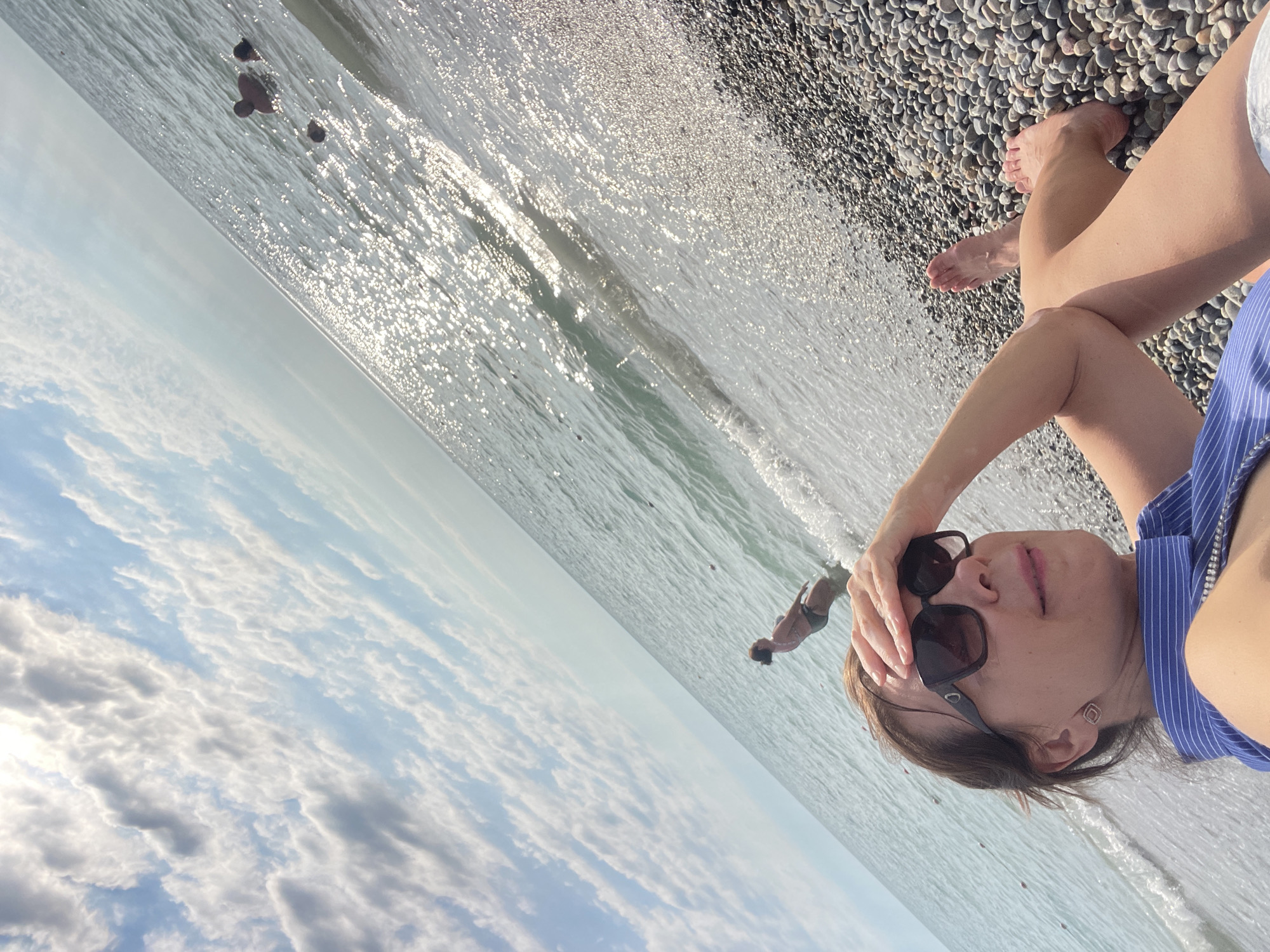 The image is a selfie taken from a low angle, with the photographer lying on a pebbled beach. The sea is in the background, with the sun reflecting off the water. The sky above is filled with fluffy white clouds.
In the foreground, the photographer is wearing sunglasses and a blue and white striped top. Their hand is resting on their head. Their legs are extended, and their feet are visible. The beach is covered in smooth, grey pebbles.
In the background, there are a few people in the water. One person appears to be swimming or wading. The water is clear and shallow, with small waves lapping at the shore.
The image appears to have been taken on a sunny day, likely in the late afternoon or early evening, given the angle of the sun and the warm lighting. The overall mood of the image is relaxed and summery. The location context is Sochi, Russia. There is no visible text in the image.