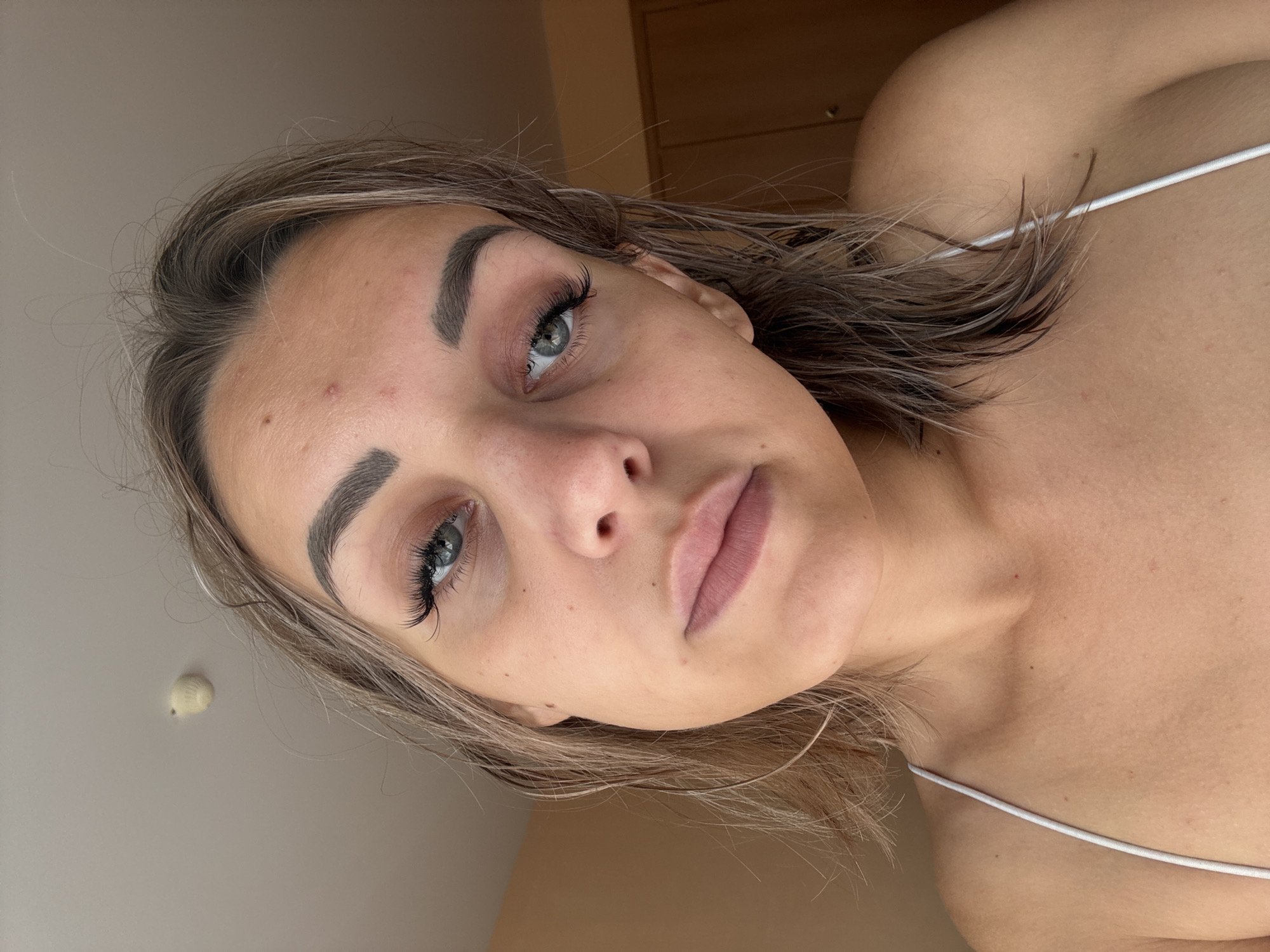 A young woman with light brown hair and blue-green eyes is captured in a close-up selfie. She is wearing a white tank top, and her hair appears to be wet and styled loosely around her shoulders. Her makeup includes noticeable false eyelashes and lip gloss. The background is a neutral-toned wall and a glimpse of a closet door. A small, light-colored object is visible on the ceiling in the upper left corner of the frame. The overall lighting suggests an indoor setting, possibly a bedroom or bathroom, during the daytime. The image does not contain any information that could indicate its location in Kholmsk, Russia.