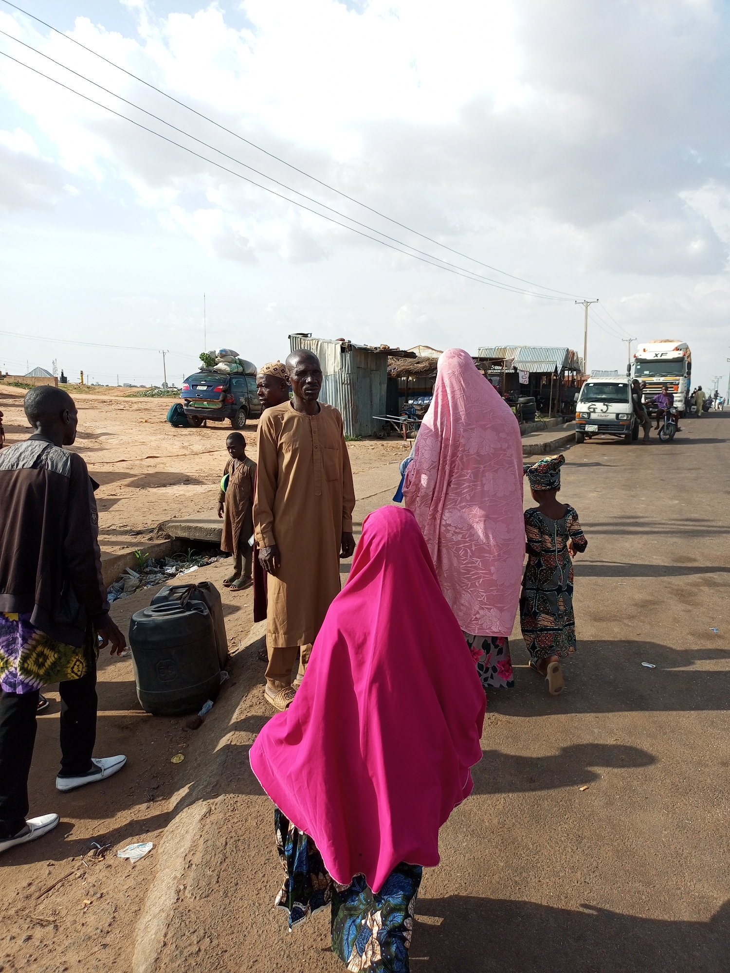 In Gadao, Nigeria, a group of people are gathered near a road. A man in a brown traditional robe stands on the right, facing the viewer, with a young boy in a similar robe beside him. To the left, two individuals are seen from the back, one wearing a brown jacket and patterned fabric, the other partially obscured. In the foreground, a person in a bright pink headscarf is facing away from the camera. Further in the background, two women in traditional attire are walking away, one in a pink head covering and the other in a patterned dress and headscarf. A small child in a colorful dress walks alongside them.
The environment is dusty, with a dirt path running alongside the paved road. Several vehicles are visible, including a dark blue SUV with luggage on top, a white truck, and a smaller white vehicle. In the middle ground, there are makeshift structures with corrugated metal roofs, suggesting a market or roadside stalls. Large black plastic containers are placed on the side of the path. Overhead, power lines stretch across the sky, which is partly cloudy. The scene appears to be an outdoor gathering or transit point.