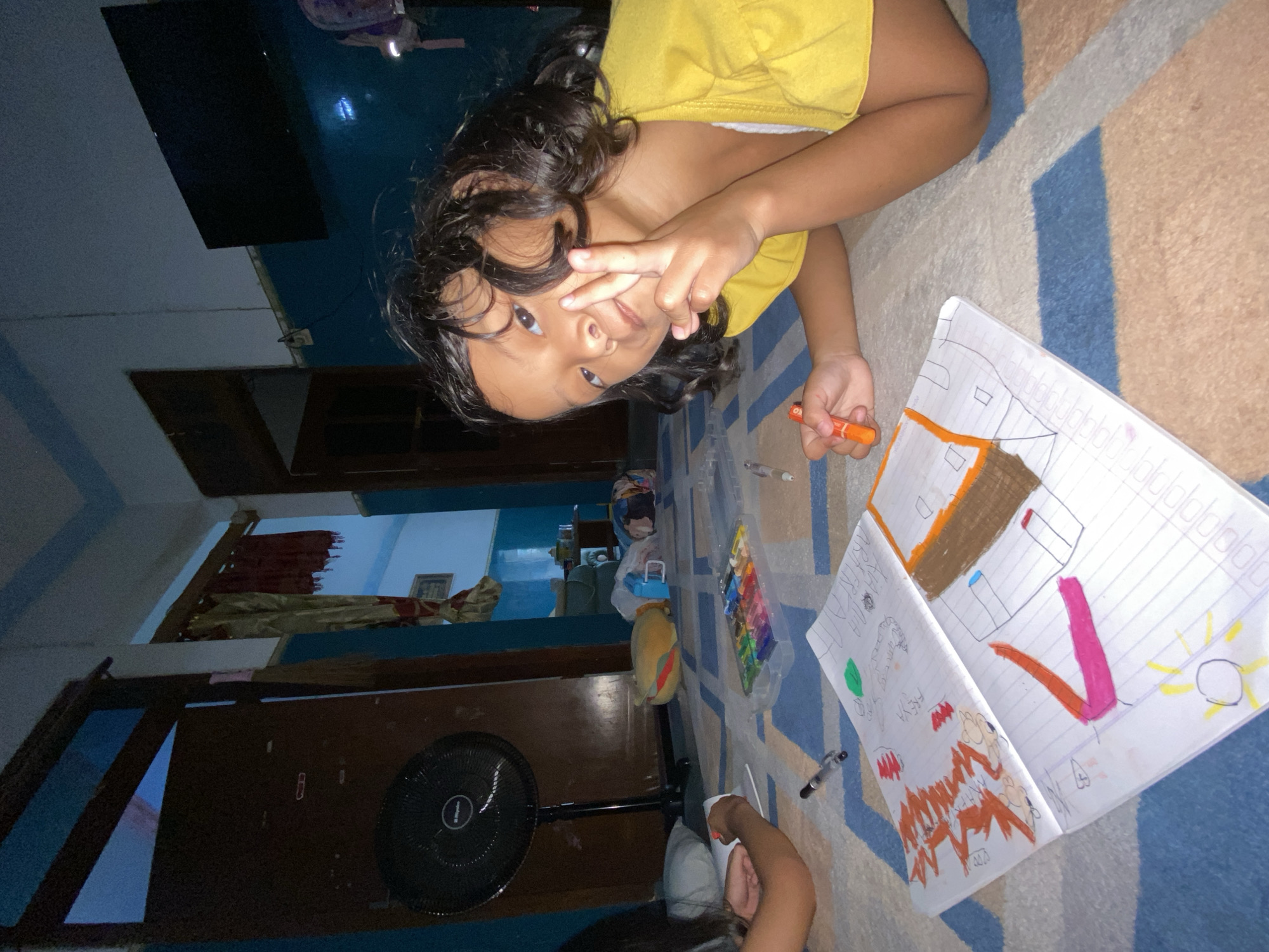 An indoor scene in Gelam, Indonesia, depicts two individuals lying on a patterned rug engaged in drawing activities. In the foreground right, a young child with dark, curly hair, wearing a yellow top and white undershirt, lies on their stomach, facing forward. The child makes a V-sign with their raised right hand and holds an orange marker in their left hand. In front of this child, an open notebook displays colorful drawings including a house with a brown roof, abstract shapes in orange and pink, a yellow sun, and red/orange scribbles. To the left, a second individual is partially visible, also lying on their stomach, drawing in a separate notebook. Between the two, a clear plastic container holds an assortment of wax crayons or oil pastels, alongside a silver pen. The rug features a geometric pattern of light brown, light blue, and darker blue. The walls are painted a dark teal or blue in the lower section, with lighter blue or white above. In the mid-ground left, a black Miyako standing fan is positioned in front of a dark brown wooden door. Further back, a large yellow plush toy and other items are on the floor. To the mid-ground right, another dark wooden door frame reveals a cream-colored curtain with a red pattern and a separate red curtain. A black flat-screen television is mounted on the upper right wall. The room is artificially lit.