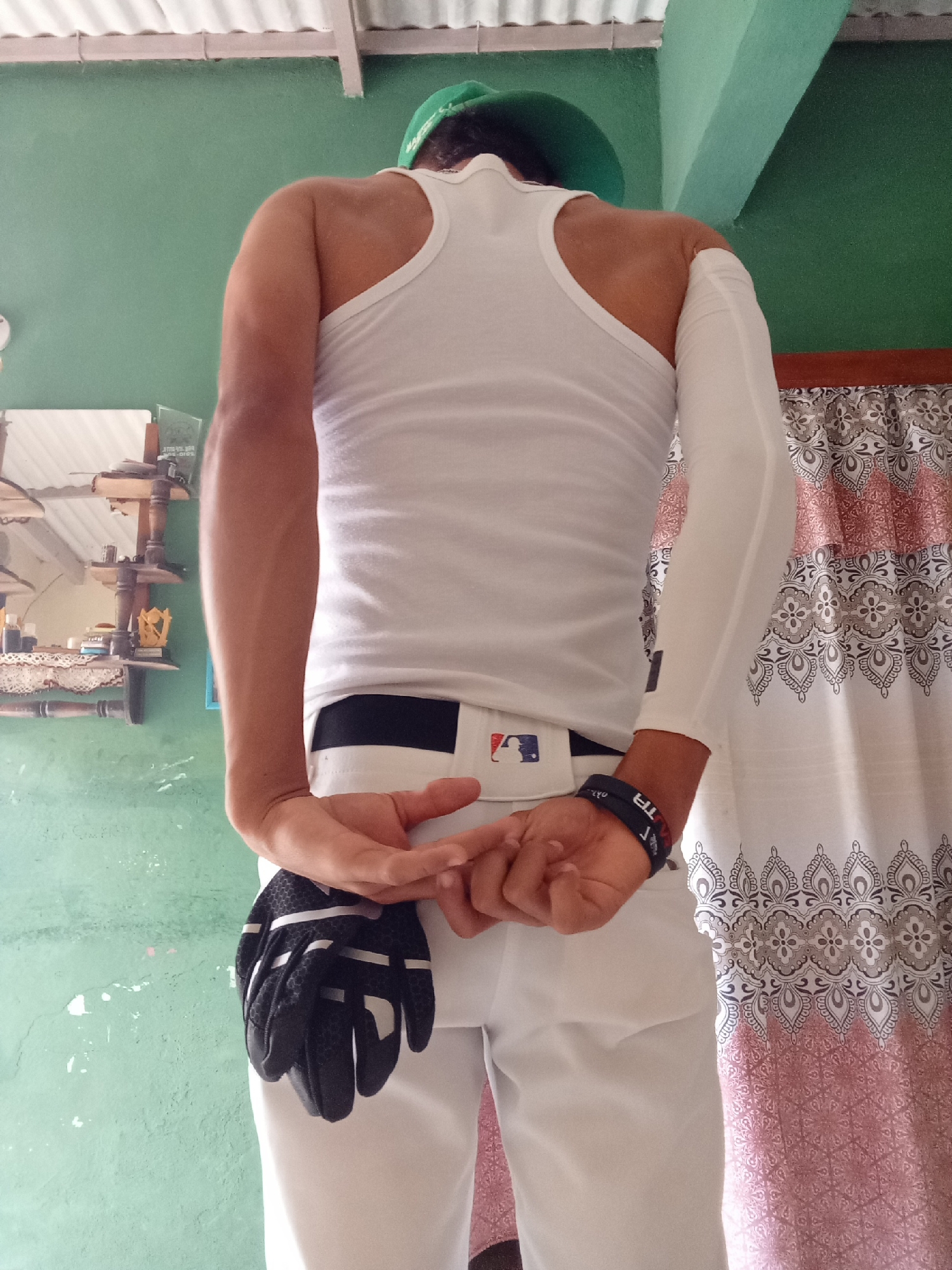A man stands with his back to the viewer, facing a green wall with visible wear and tear. He is wearing a white tank top, white pants, and a white belt with a dark logo. The man has a green object on his head, possibly a hat. A white sleeve covers his right arm. He holds a black baseball glove behind his back, his hands interlocked. A dark wristband is visible on his left wrist. To the left, a mirror reflects a shelf with decorative items. To the right, a curtain with a geometric pattern hangs. There is no specific indicator of the location within the city of Juan Griego, Venezuela.