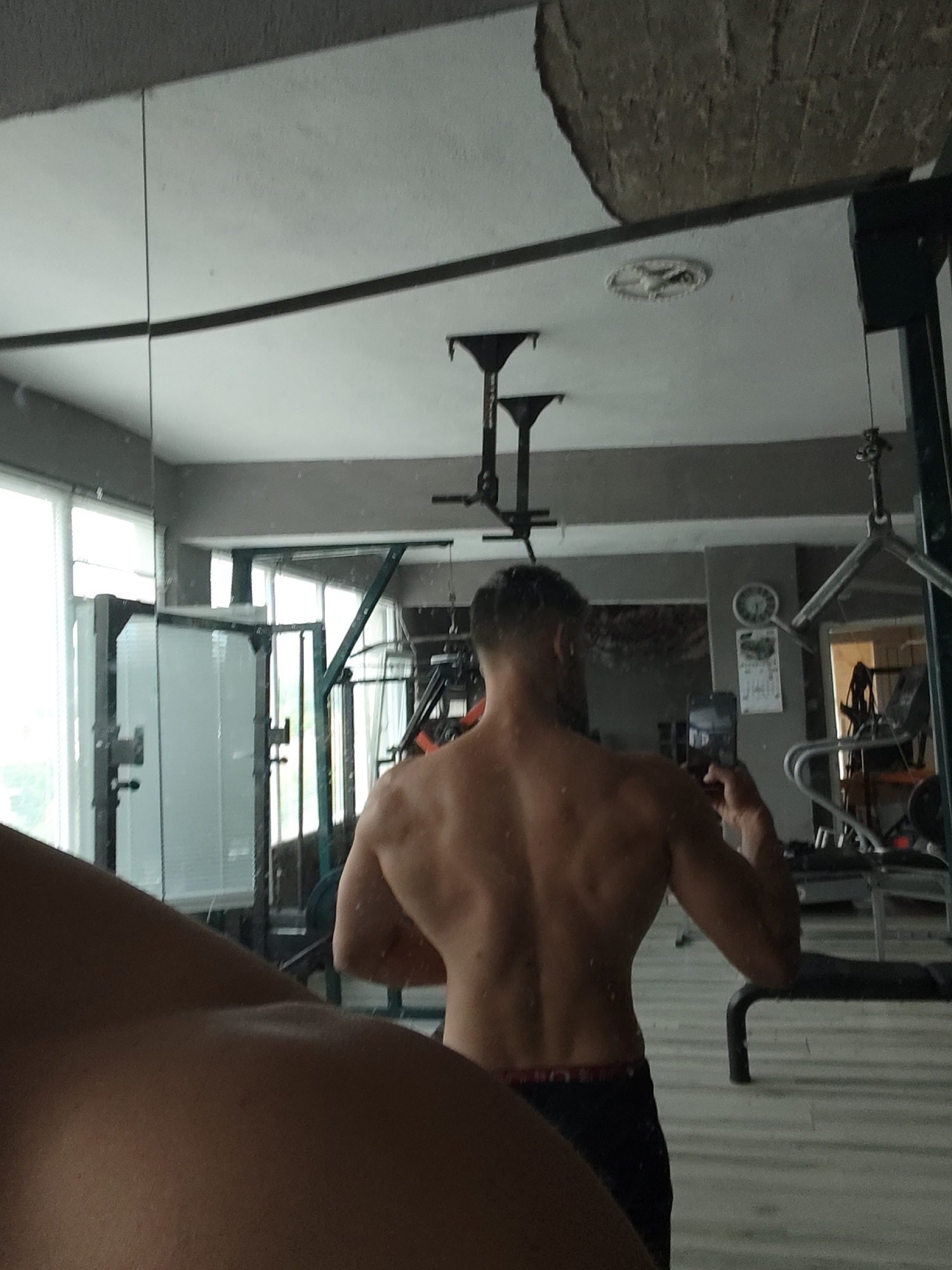 A man stands shirtless in what appears to be a gym, taking a photo of his back muscles in a mirror. Various gym equipment is visible in the reflection, along with windows. The location appears to be indoors. The city is unknown, and the state is unknown.