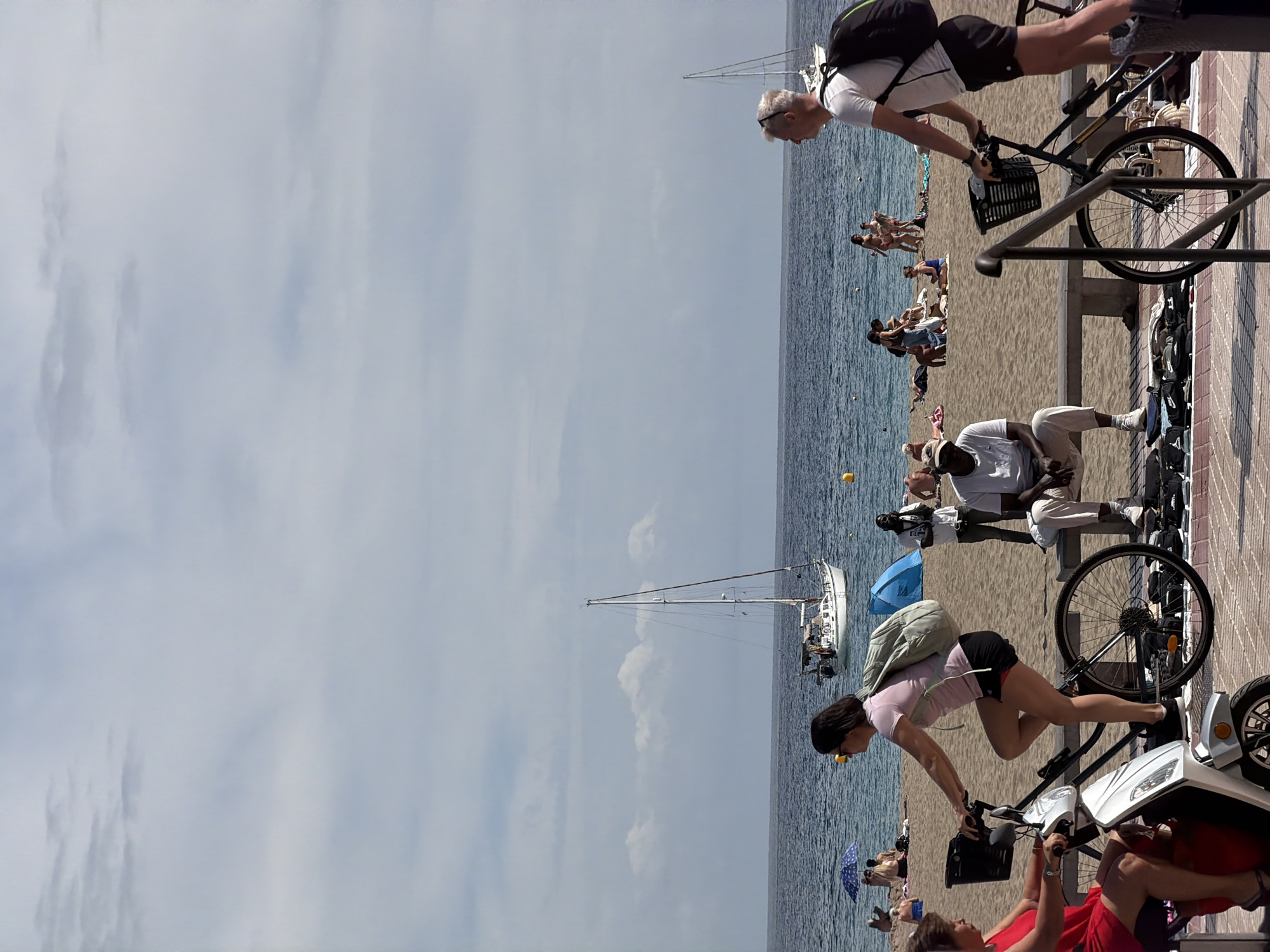 This image captures a vibrant, sunny day at a beach and promenade in Los Cristianos, Spain. The scene is horizontally divided into three main sections: the ocean on the left, a sandy beach in the middle, and a paved promenade on the right.
In the foreground on the promenade, several people are engaged in leisure activities. A woman in a light pink top and dark shorts, carrying a light green backpack, leans over a bicycle. Her body is in a dynamic, leaning pose. Another person in a red top is visible further right, appearing to interact with a white scooter or moped. A man in a white shirt and light pants stands near another bicycle on the promenade.
Further along the promenade, another man with a dark backpack and white t-shirt is seen riding a bicycle. The promenade features dark metal railings and a patterned paved surface.
The middle ground is occupied by the sandy beach, where numerous people are relaxing, sunbathing, or swimming in the ocean. Some individuals are under colorful beach umbrellas, including a blue one and one with white polka dots. Small yellow buoys are scattered across the calm blue water.
In the background, the vast expanse of the ocean stretches towards the horizon under a light blue sky dotted with scattered white clouds. A white sailboat with its mast prominent is anchored or sailing in the distance. The bright sunlight suggests it is midday or early afternoon, contributing to the warm and leisurely atmosphere of the scene. No discernible text is visible in the image.