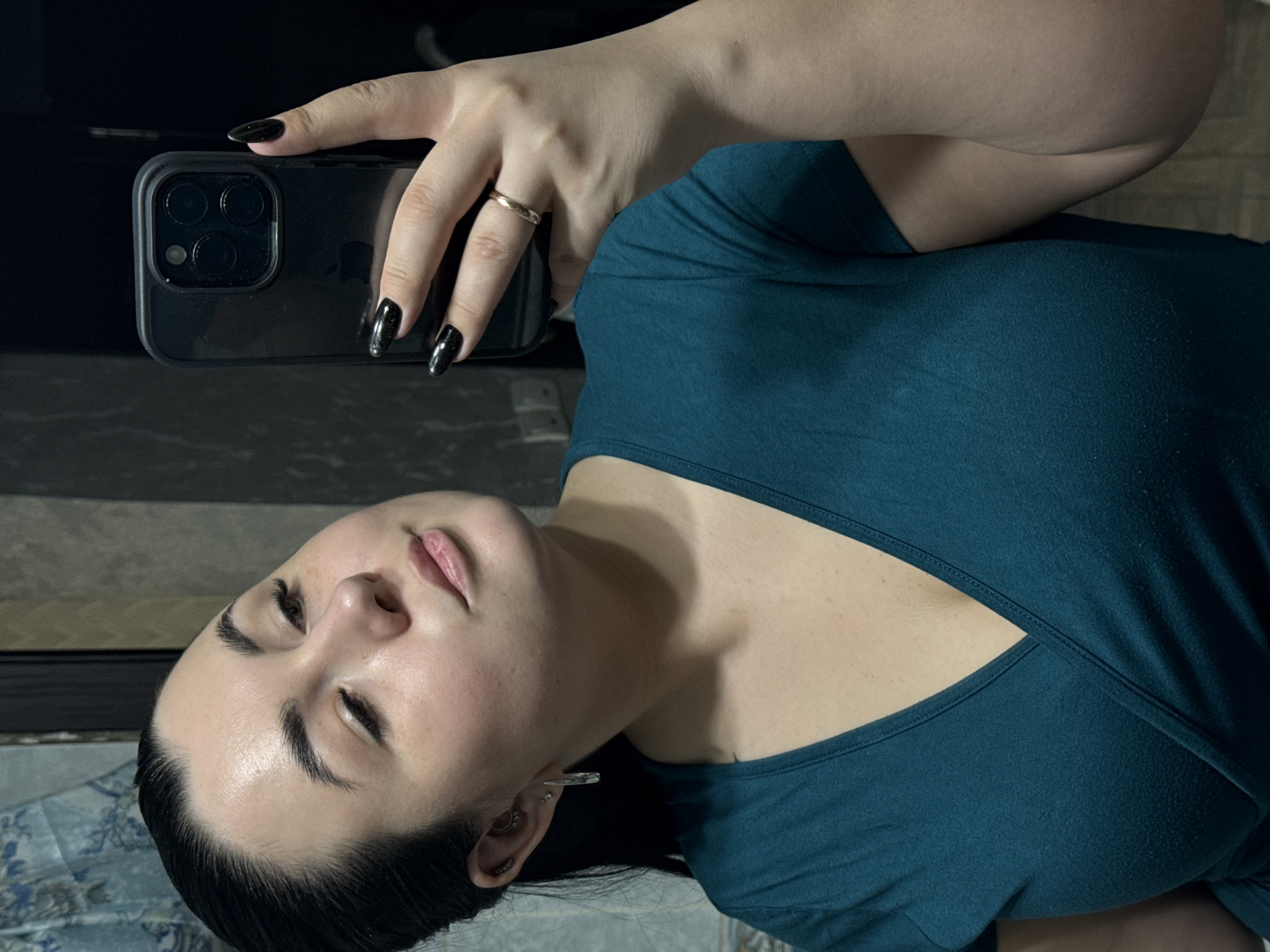 A woman takes a selfie with a black iPhone. She has fair skin, dark hair, and is wearing a teal v-neck shirt and a gold ring. Her fingernails are painted black. She is wearing multiple earrings. The background is blurry and does not clearly depict a specific location in Chaikovsky, Russia. There is no other information that indicates the location is in Chaikovsky, Russia.