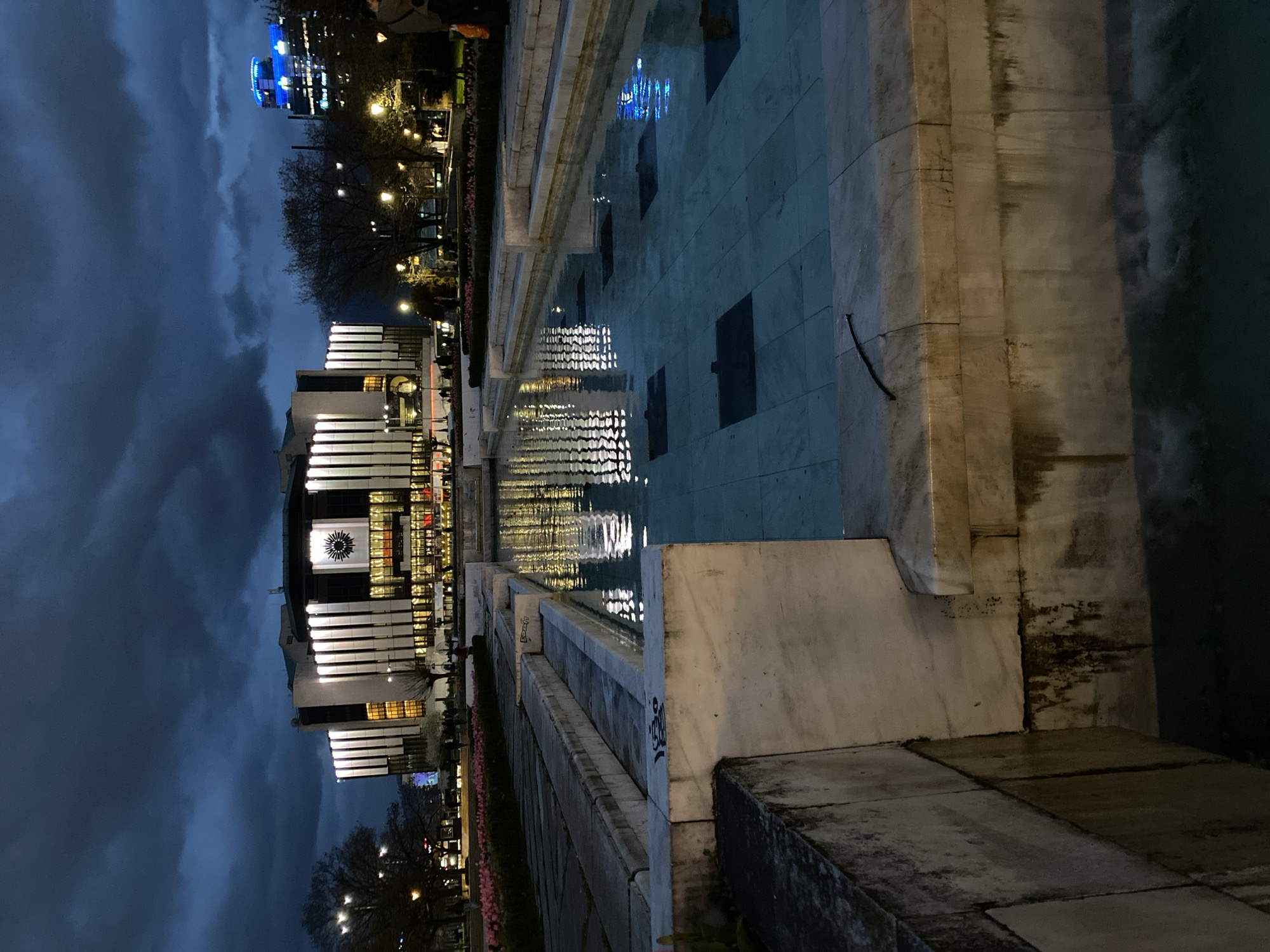 The image captures the National Palace of Culture in Sofia, Bulgaria, reflected in a water feature. The architecture of the palace is characterized by its elongated horizontal lines and illuminated windows. The sky is dark and cloudy. The water reflects the palace and the city lights, creating a rippled effect. A tiled surface surrounds the water feature, with a stone wall along the edge, with some graffiti. Trees and other lights are visible in the background, indicating an urban setting. No people are clearly visible in the immediate foreground.