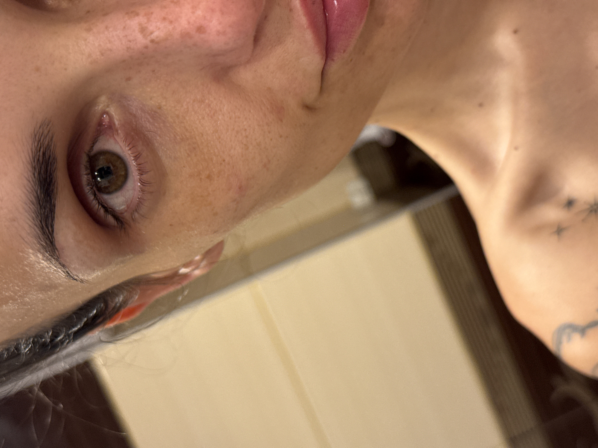 A close-up, slightly angled photograph depicts a portion of a woman's face, specifically the left eye, cheek, and part of her neck and collarbone. The eye is brown with visible eyelashes and eyebrows. Freckles are present on the cheek. Her lips appear to have a glossy finish. A tattoo is visible on the collarbone area. The background is out of focus and appears to be a light-colored surface with vertical lines. No information about the location or Pleven, Bulgaria is discernible from the image.