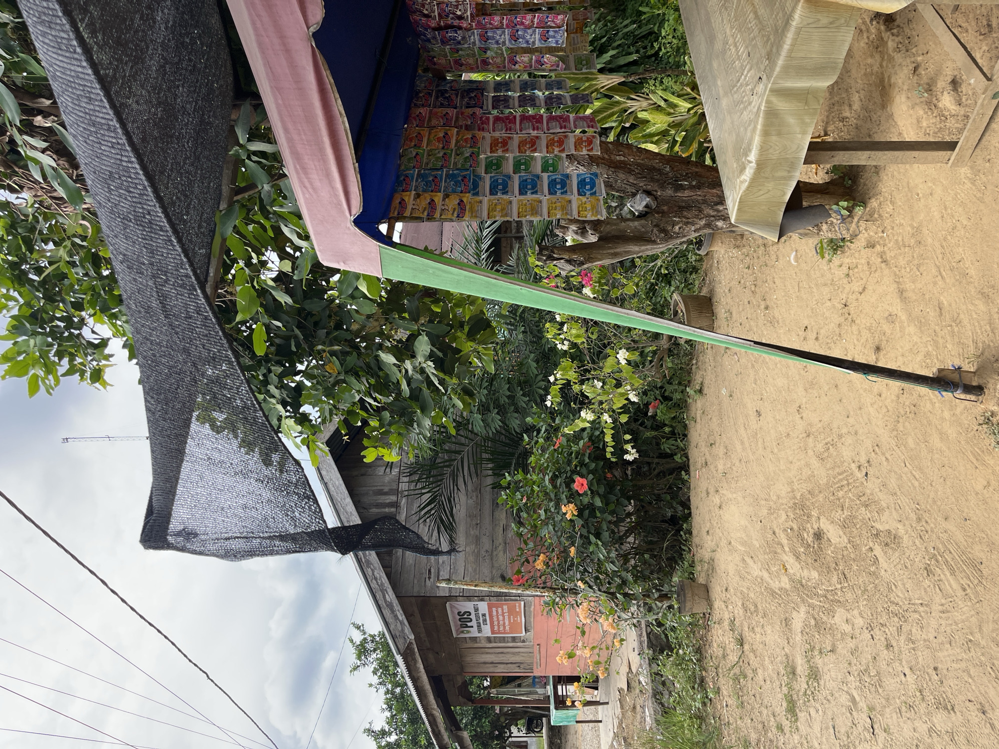 The image depicts a roadside scene in Sukajadi, Indonesia, featuring a small wooden building with a sign indicating "POS" (Post Office) and "PERKASA PAKET C SEKITAR RIAU." In front of the building, there is a display of colorful packaged goods hanging under a canopy. To the right, a covered table sits on a sandy ground. The environment includes lush green foliage, blooming flowers, and a general sense of a rural or semi-urban setting. The sky is overcast. No people are visible in the frame.