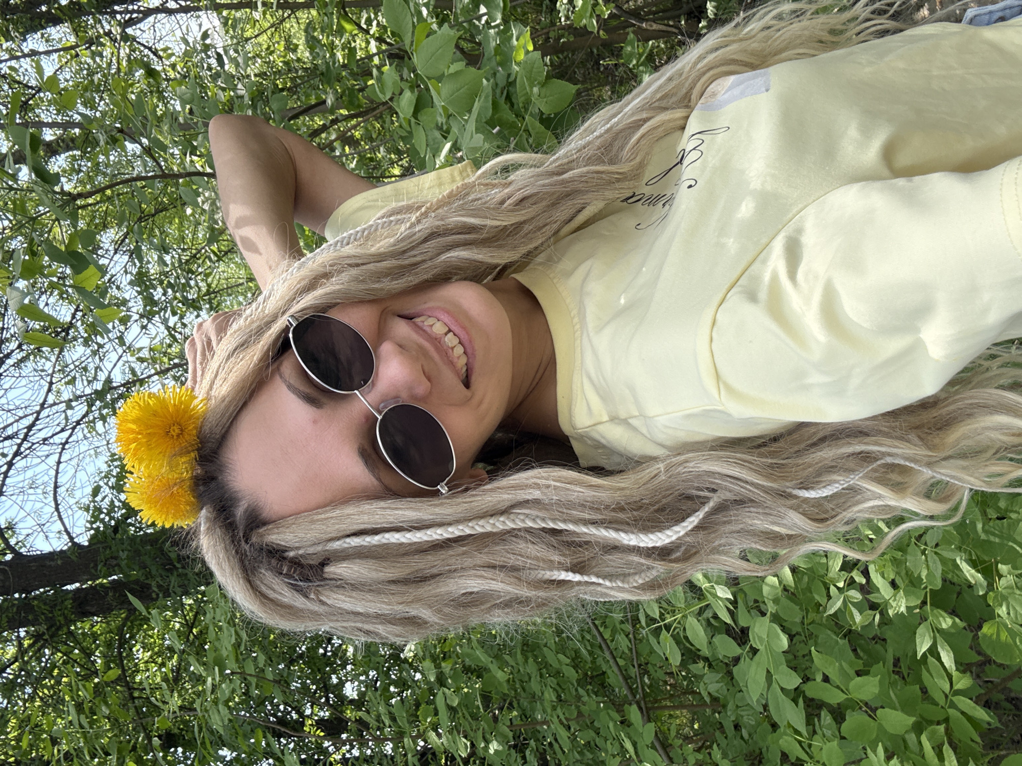 A young woman with braided hair, wearing round sunglasses and a yellow t-shirt, lies on her back in a grassy area surrounded by green foliage. She has a yellow dandelion tucked behind her ear. The setting appears to be outdoors, possibly a park or garden. Location: Rusanovo, Russia.