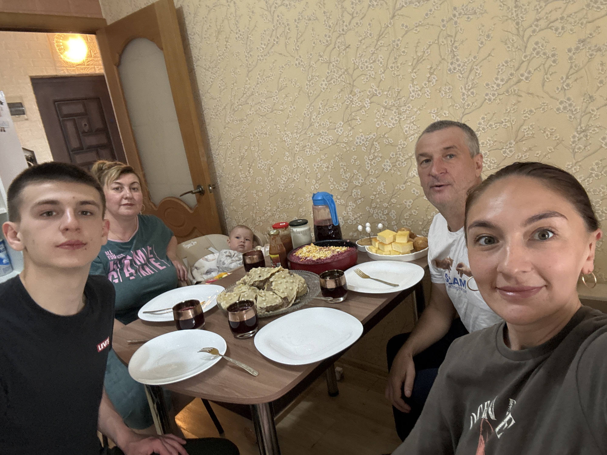 A group of six people are gathered around a table for a meal. A young man is positioned to the left, wearing a black t-shirt, while a woman in a green t-shirt sits next to him. A baby sits in a high chair at the table's edge. Across the table, a man wearing a white t-shirt sits to the right, and a woman in a gray-green t-shirt holds a phone up to take the picture.
The table is set with white plates, gold-colored forks, and glasses containing a dark liquid. Various dishes are arranged on the table, including what appears to be sliced bread, a salad or dip in a red bowl, and yellow squares stacked on a plate. A juice jug and other food items are also present.
The room has patterned wallpaper, and a wooden door is visible in the background. Based on the style of interior design and the people's clothing, the image appears to be taken in someone's home. There's no information available in the picture about the location being in the city of Sochi, Russia.