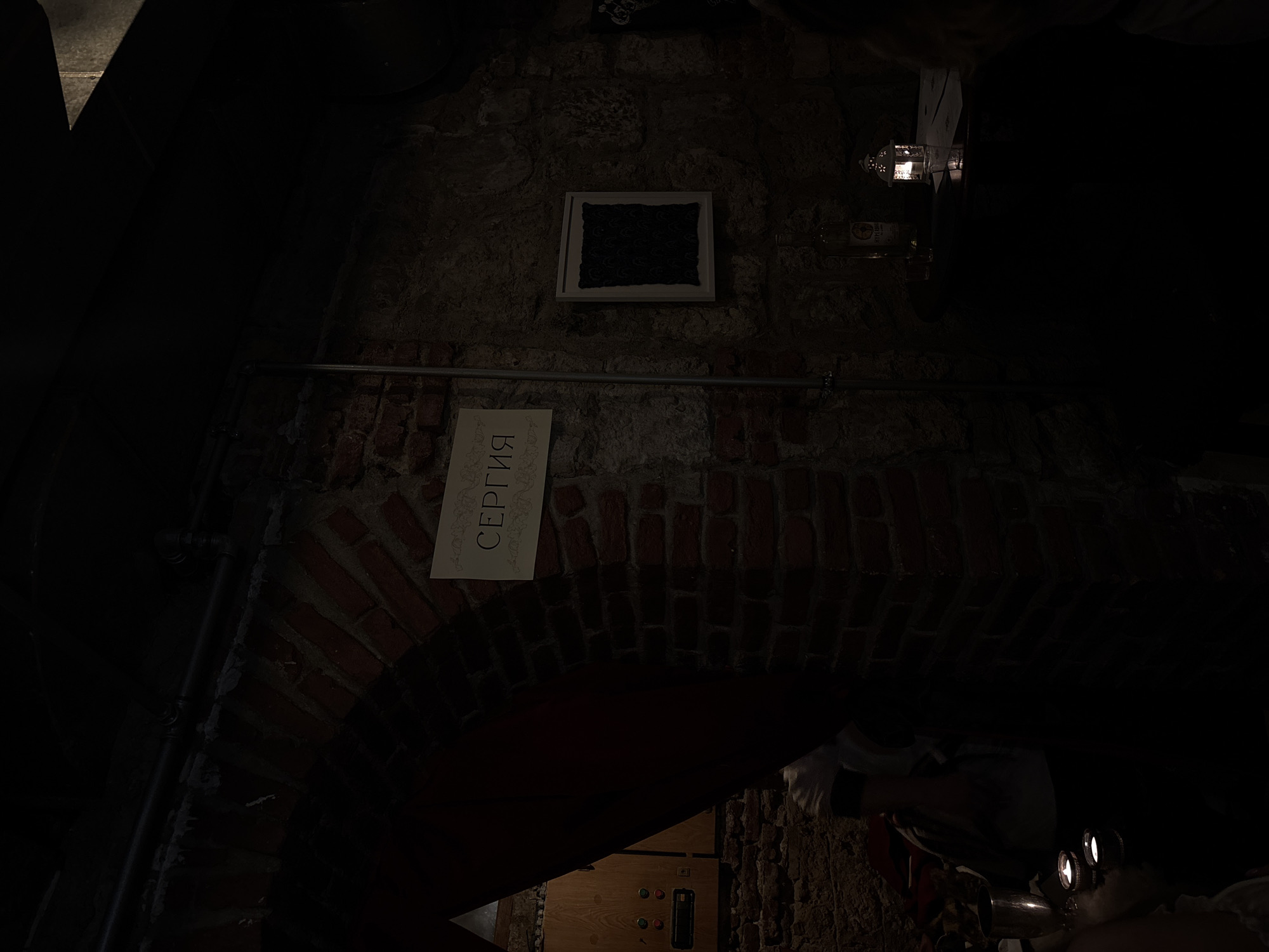 The image shows a dimly lit interior, possibly a restaurant or bar in Varna, Bulgaria, based on the Cyrillic script on a sign that reads "Сергия" (Sergiya). The sign is mounted on a brick archway. Above the sign, there's a framed, dark textile or patterned artwork on the wall. A metal pipe runs horizontally across the brick wall, supporting the sign. Below the archway, there is a wooden panel with buttons and a narrow slot, possibly part of a control panel. At the right edge of the frame, there are glimpses of people, including white sleeves, suggesting service staff. There are also lights, possibly spotlights or headlamps, which contribute to the dim lighting. The overall environment suggests a rustic or vintage aesthetic, possibly within a basement or cellar space.