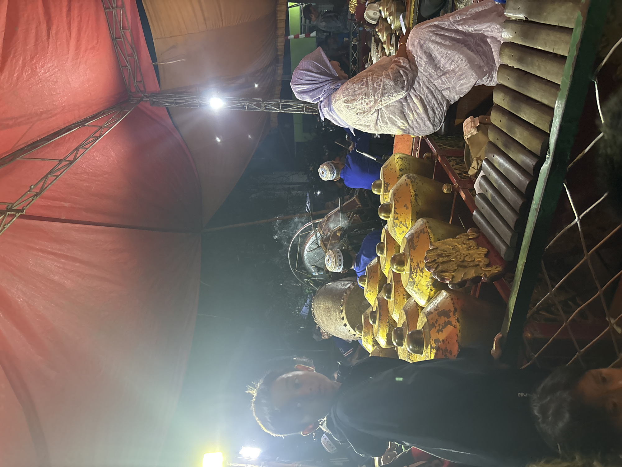 The image depicts a gamelan performance in Panongan, Indonesia, under a red tent. In the foreground, a young boy in a black hoodie looks towards the camera. To his right, a woman in a light purple hijab and patterned dress is seated, her hands resting on a wooden xylophone-like instrument. Behind them, a set of large, golden-yellow kettle gongs, typical of a gamelan orchestra, are arranged. Further back, other musicians are visible, some wearing white skullcaps, playing various instruments including a large drum. The background is dimly lit, showing glimpses of trees and buildings. The overall scene suggests an outdoor cultural event or performance.