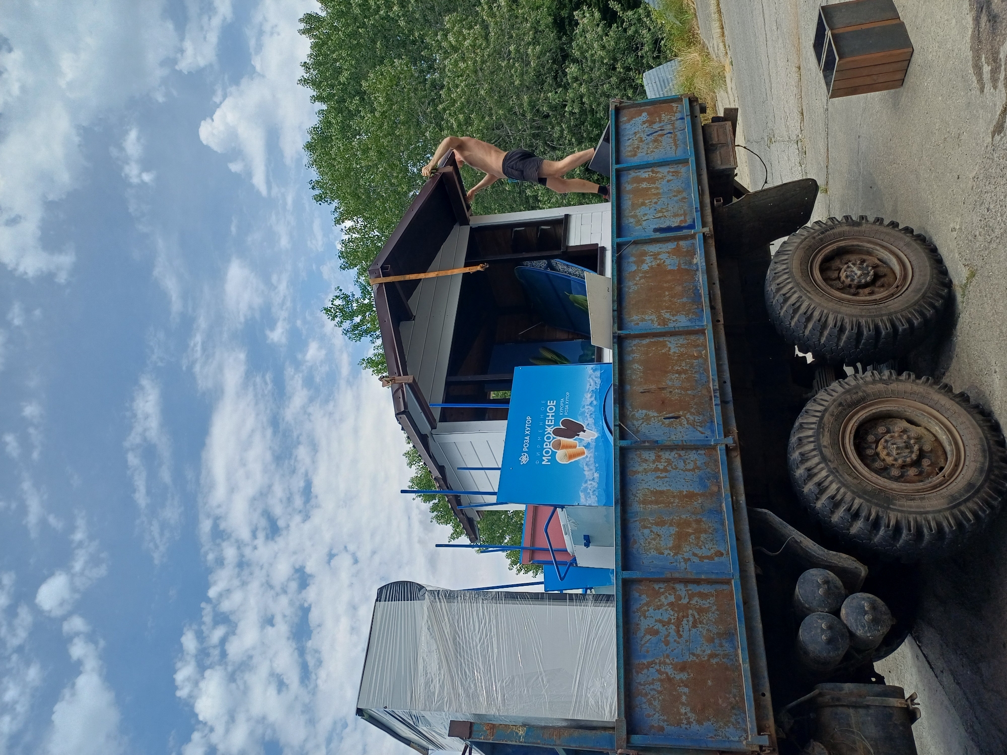 A shirtless person wearing black shorts and red socks stands on the back of a rusty, blue flatbed truck and appears to be adjusting the roof of a small, white structure. The structure is loaded onto the truck bed, alongside other items, including a large white appliance wrapped in plastic and a blue sign with text and images of ice cream. The truck has large, rugged tires. In the background, lush green trees are visible against a cloudy blue sky. To the right of the truck, two wooden planters sit on the ground. The scene is outdoors, on a paved surface.