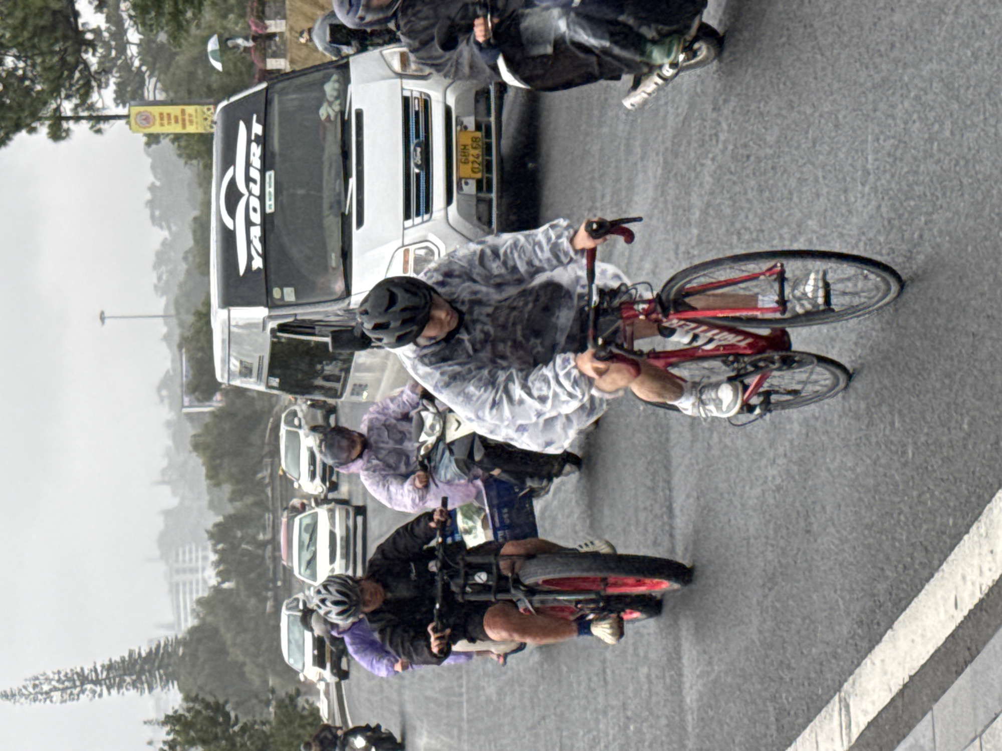In Da Lat, Vietnam, on a rainy day, a cyclist wearing a clear plastic poncho and a black helmet rides a red bicycle with thick tires on a wet asphalt road. He is in the foreground, with his hands on the handlebars and his feet on the pedals. Behind him, a person on a scooter with a purple poncho is visible. Further back, a white van with "YAQURT" written on the front is driving. Several other vehicles, including cars and motorcycles, are on the road, all appearing to navigate the wet conditions. The environment suggests an urban setting with trees and buildings in the background, shrouded in the overcast weather. The road surface is visibly wet and reflective.