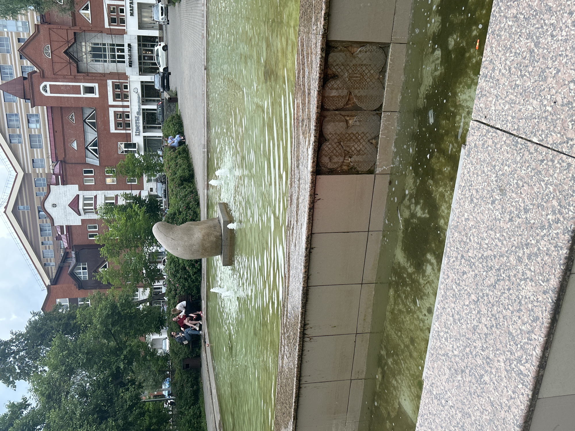 The image depicts an outdoor scene featuring a fountain and adjacent buildings, likely in Perm, Russia. The fountain's water is a greenish hue and appears somewhat murky, with ripples suggesting movement and small jets of water visible. In the foreground, a large, abstract stone sculpture with a curved, smooth form emerges from the water. The fountain's edge is lined with textured granite and tiled surfaces.
In the background, several people are seated on a bench near the fountain, some appearing to be interacting or relaxing. Lush green trees and bushes surround the area, providing a natural element to the urban setting. Behind the greenery, multi-story buildings with red brick and white architectural details are visible, suggesting a commercial or residential area. One building has prominent windows and decorative elements.
The overall impression is that of a public space, possibly a park or plaza, during daylight hours. The weather appears overcast, contributing to a somewhat muted light. The scene suggests a moment of leisure, with people enjoying the fountain area. There is no discernible text in the image that provides specific information beyond what is visually present in the architecture.