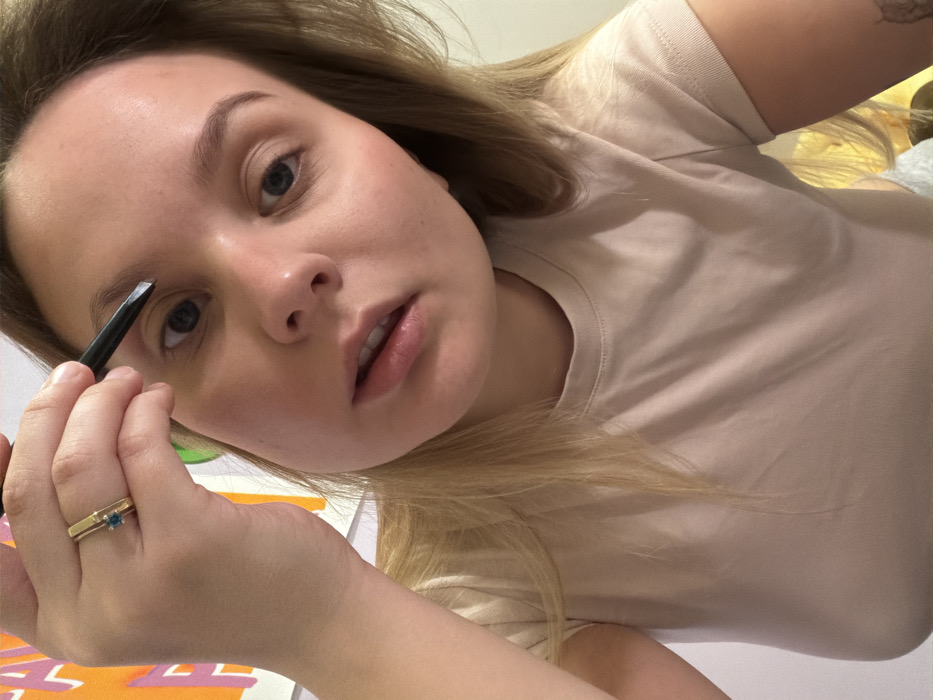 A young woman with light brown hair and light blue eyes is captured in a close-up, slightly angled shot. She is wearing a plain light beige t-shirt and is focused on applying makeup. Her left hand holds a dark eyebrow pencil or brush, which she is using to define or fill in her left eyebrow. A gold-toned ring with a small blue stone adorns her left ring finger. Her gaze is directed intently, likely at a mirror or the camera she's using for the shot, with a neutral and concentrated expression. The background is softly blurred, showing vague outlines of colorful shapes, suggesting an indoor setting. The lighting is soft and even, indicating an indoor environment. There is no visible text within the image. The scene depicts a moment of personal grooming in Tel-Aviv, Israel.