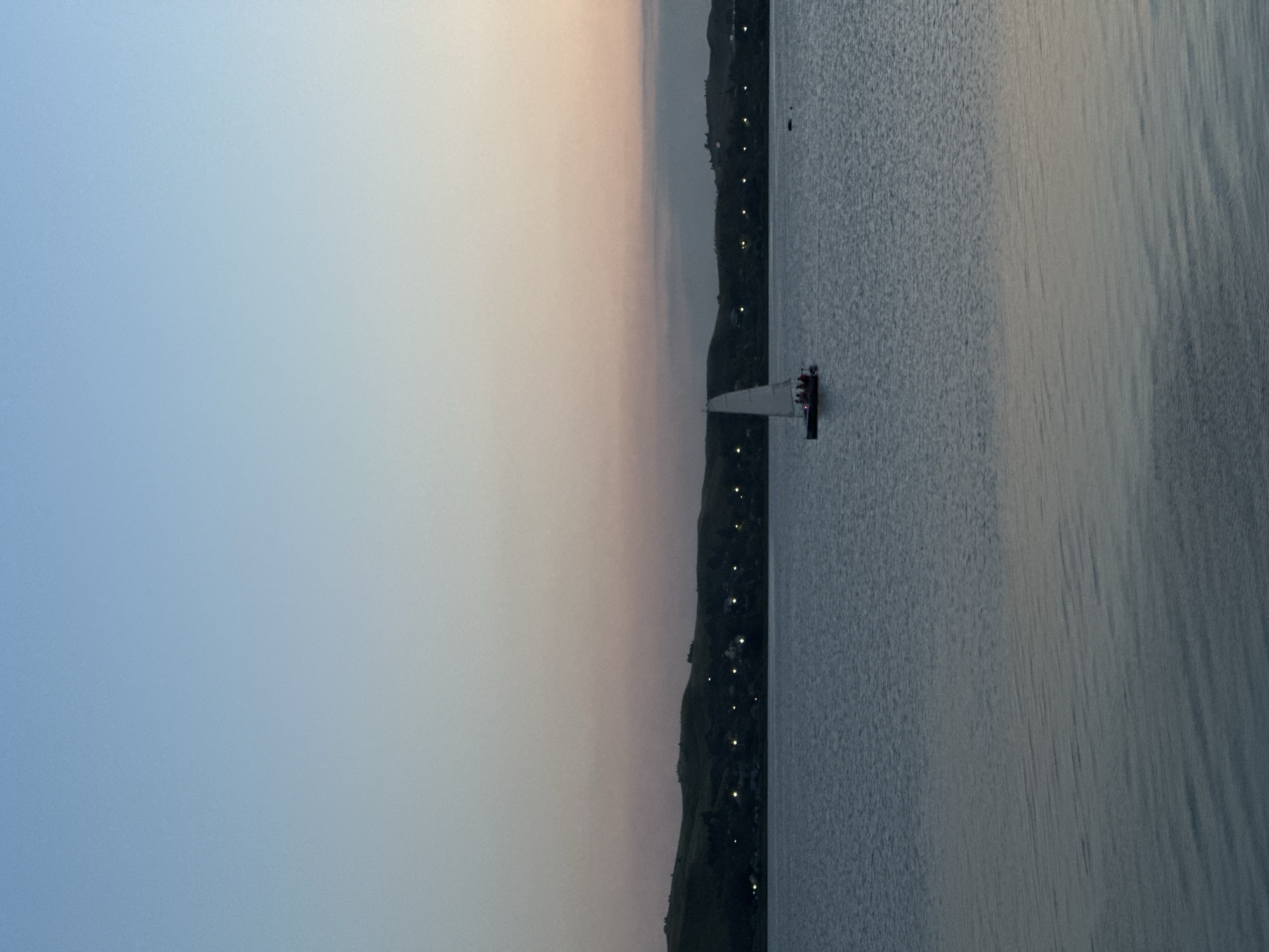 An aerial view captures a serene lake scene at dusk, possibly located near Kokshetau, Kazakhstan. The sky transitions from a clear, pale blue at the top to soft hues of pink and orange near the horizon. Below the sky, a line of rolling, dark hills forms the backdrop, dotted with small, illuminated lights that suggest a settlement. The vast expanse of the lake stretches across the foreground, its surface rippling gently. In the middle distance, a small sailboat navigates the water, its white sail illuminated against the darkening sky. The boat appears to have several people on board. A dark, circular object, possibly a buoy or a submerged rock, is visible to the right of the sailboat. The overall atmosphere is tranquil and picturesque.