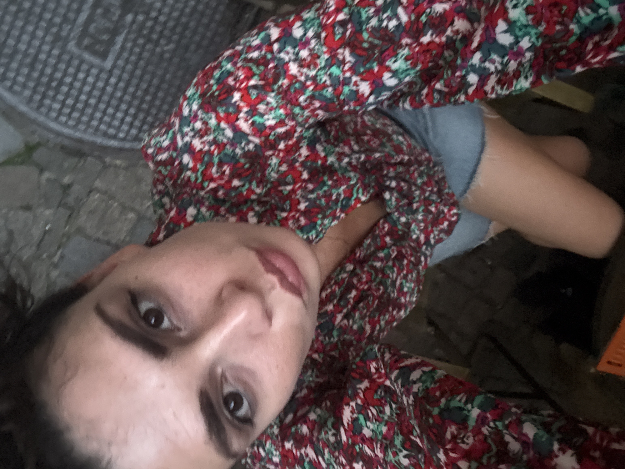 The scene, located in Sofia, Bulgaria, captures a single individual from a low-angle, upward-facing perspective. The subject, a woman with dark hair and eyes, is positioned centrally, looking directly into the camera. She wears a long-sleeved blouse adorned with a dense floral pattern in shades of red, green, and white, paired with light blue denim shorts. Her expression features a slight pout. The background consists of grey paving stones, with a circular, dark grey metal manhole cover, exhibiting a grid-like texture, visible in the upper right. A partial orange object is discernible in the lower-left corner of the frame.