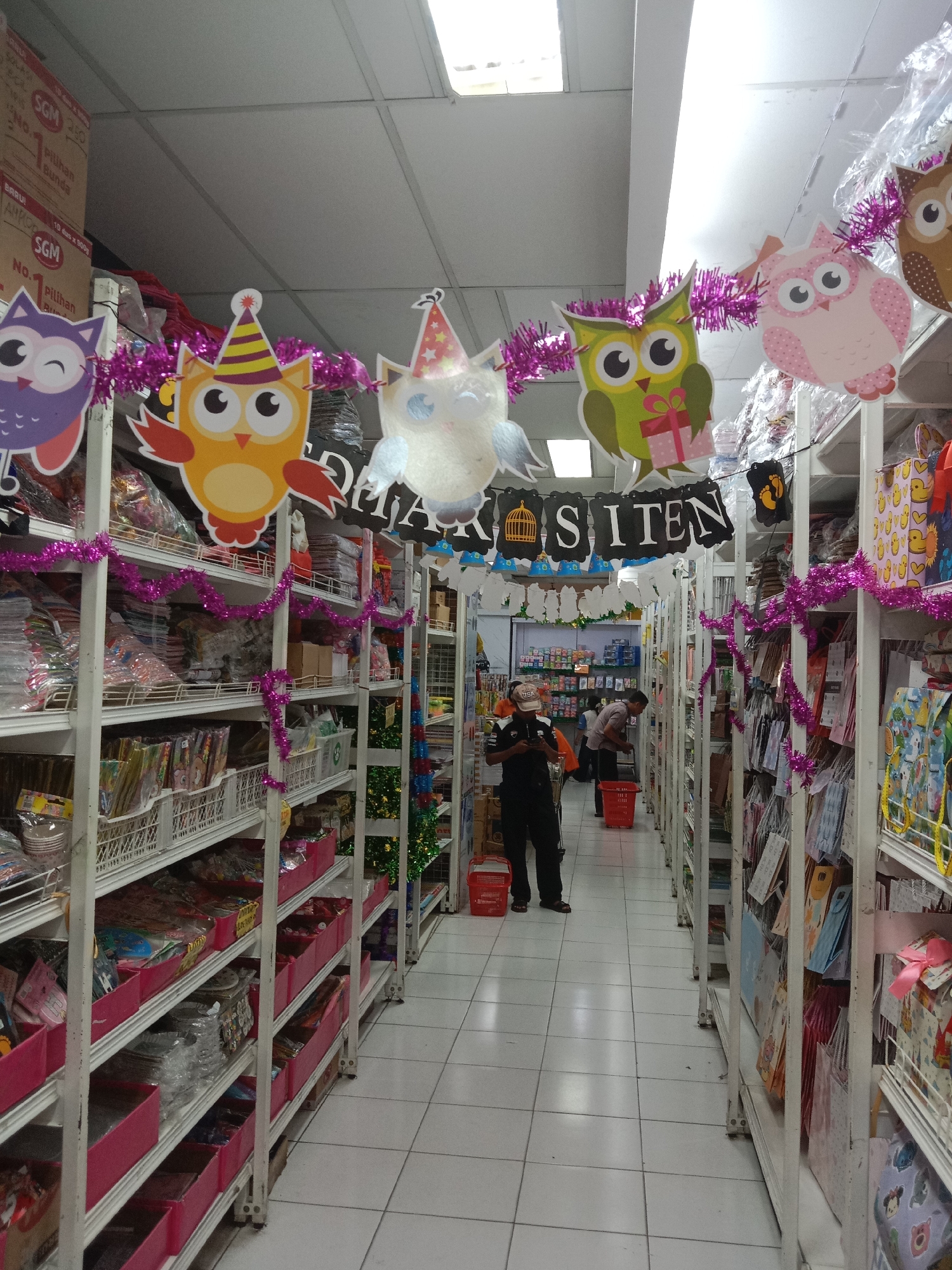 The image depicts the interior of a retail store, likely a party supply or gift shop, located in the city of Probolinggo, Indonesia. The store features tall, white shelving units filled with various products, many of which appear to be packaged party favors, gift bags, and decorations. The aisle is illuminated by overhead fluorescent lighting.
In the foreground and mid-ground, the shelves are decorated with garlands of purple tinsel and colorful owl-shaped cutouts wearing party hats, suggesting a festive theme, possibly a birthday celebration. A banner with the word "SITEN" is visible, suspended above the aisle.
Two individuals are present in the scene. One person, wearing a black uniform with an "USA" patch and a cap, stands in the left aisle, looking at a mobile device. This individual appears to be an employee or security personnel. Another person, wearing a light-colored shirt and dark pants, is browsing items on the shelves in the right aisle, holding a red shopping basket.
The floor is tiled with white square tiles. Cardboard boxes are stacked on shelves in the background, and some products are wrapped in plastic. The overall impression is a well-stocked, organized retail space decorated for a special occasion.
