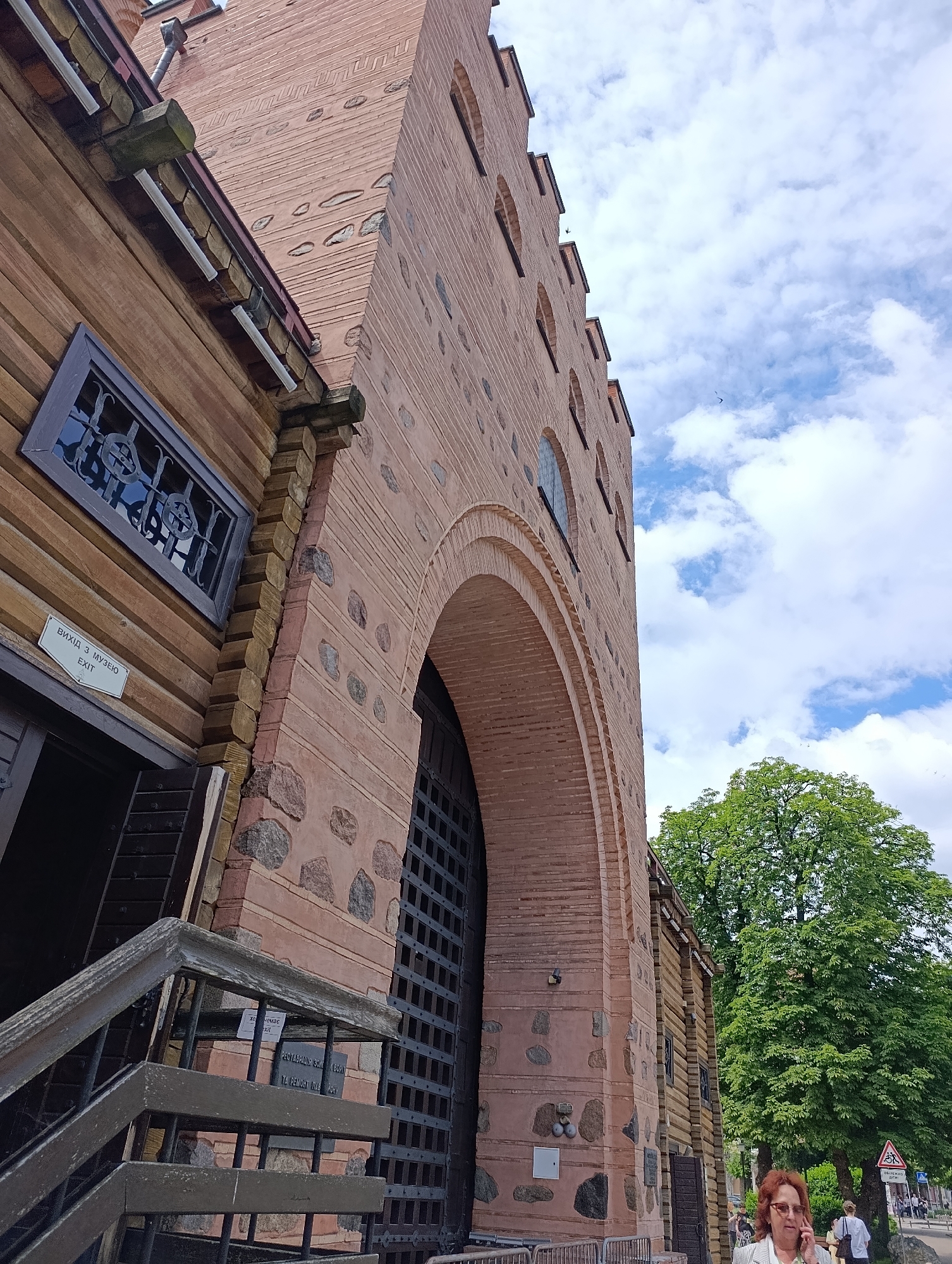 In Kyiv, Ukraine, the image shows a low-angle view of a historical building with a large arched entrance and a wooden facade. The building appears to be made of brick and stone, with several small arched windows on its upper section. A sign on the wooden part reads "ВИХІД З МУЗЕЮ" and "EXIT". To the right of the building, a large green tree is visible, and beyond that, a street scene with people and a traffic sign. One woman is prominently featured in the lower right corner, talking on a mobile phone and wearing glasses. The sky above is partly cloudy.