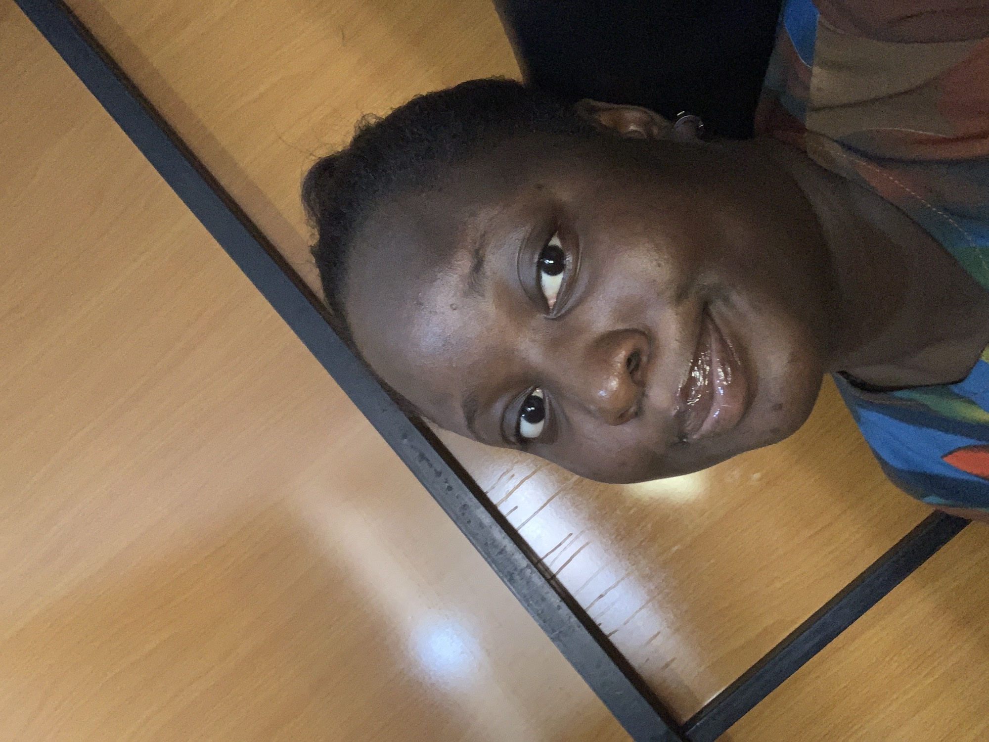 The image shows a close-up, selfie-style shot of a young Black woman. She is looking towards the camera with a slight smile and her lips are glossy. Her hair is dark and pulled back. She is wearing a multi-colored, patterned top. The background appears to be a wooden surface with a dark, angled bar across it. The lighting suggests an indoor setting. There are no discernible urban or environmental features to indicate a specific city or location.