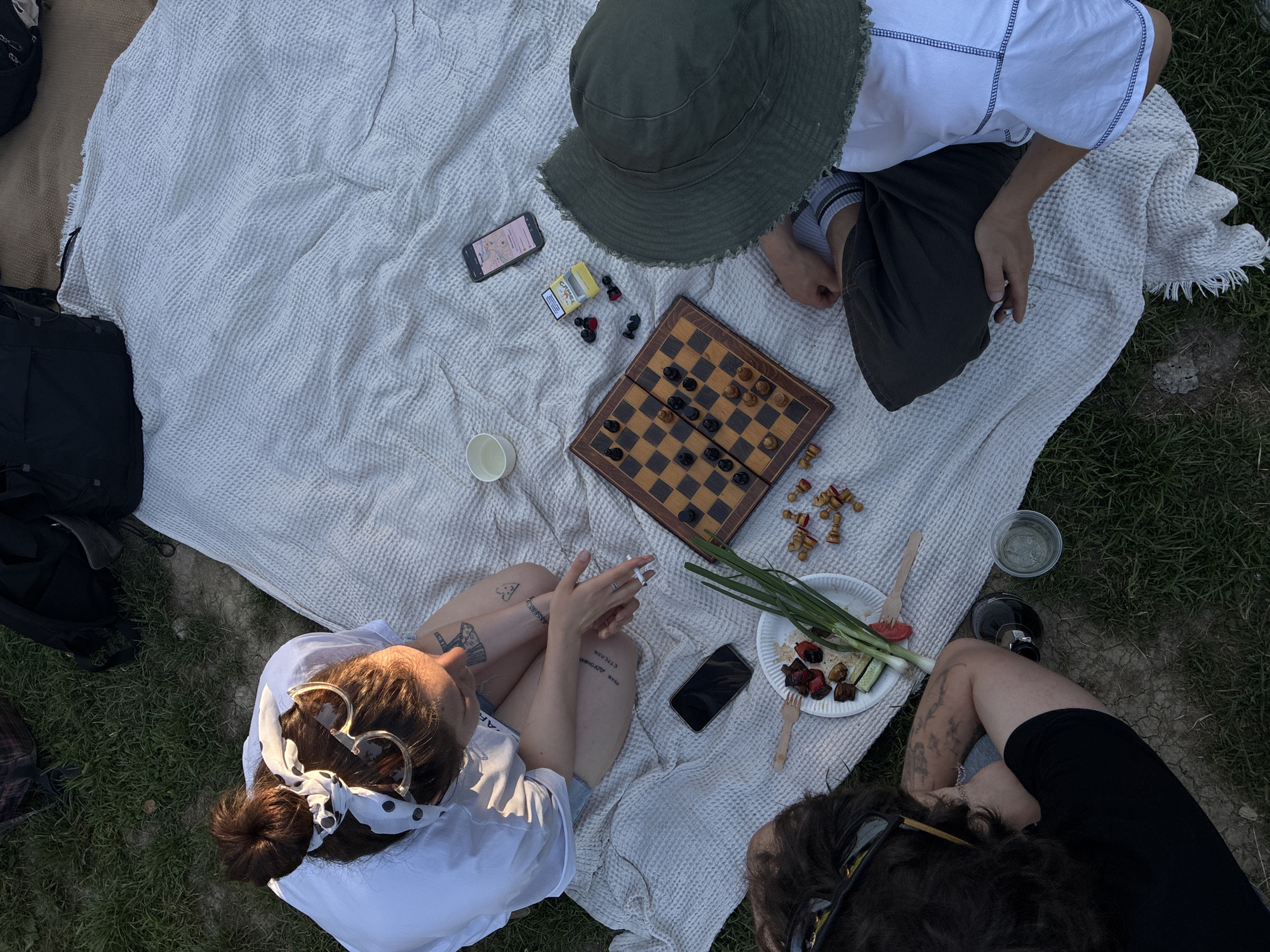 Overhead shot of a picnic scene, potentially located in the Cukarica Urban Municipality, Serbia. Four people are situated on a white blanket laid on grass. One person is wearing a white shirt and heart-shaped sunglasses with a white, patterned headband. Another person wears a white shirt and olive green bucket hat. One person is wearing dark clothing. A chess board is in the center with pieces set out. A white plate contains scallions, grilled vegetables, and a wooden fork. A cell phone lies on the blanket next to the plate, and another cell phone is adjacent to the chess board. There are other containers on the blanket, and two bags are visible in the periphery.