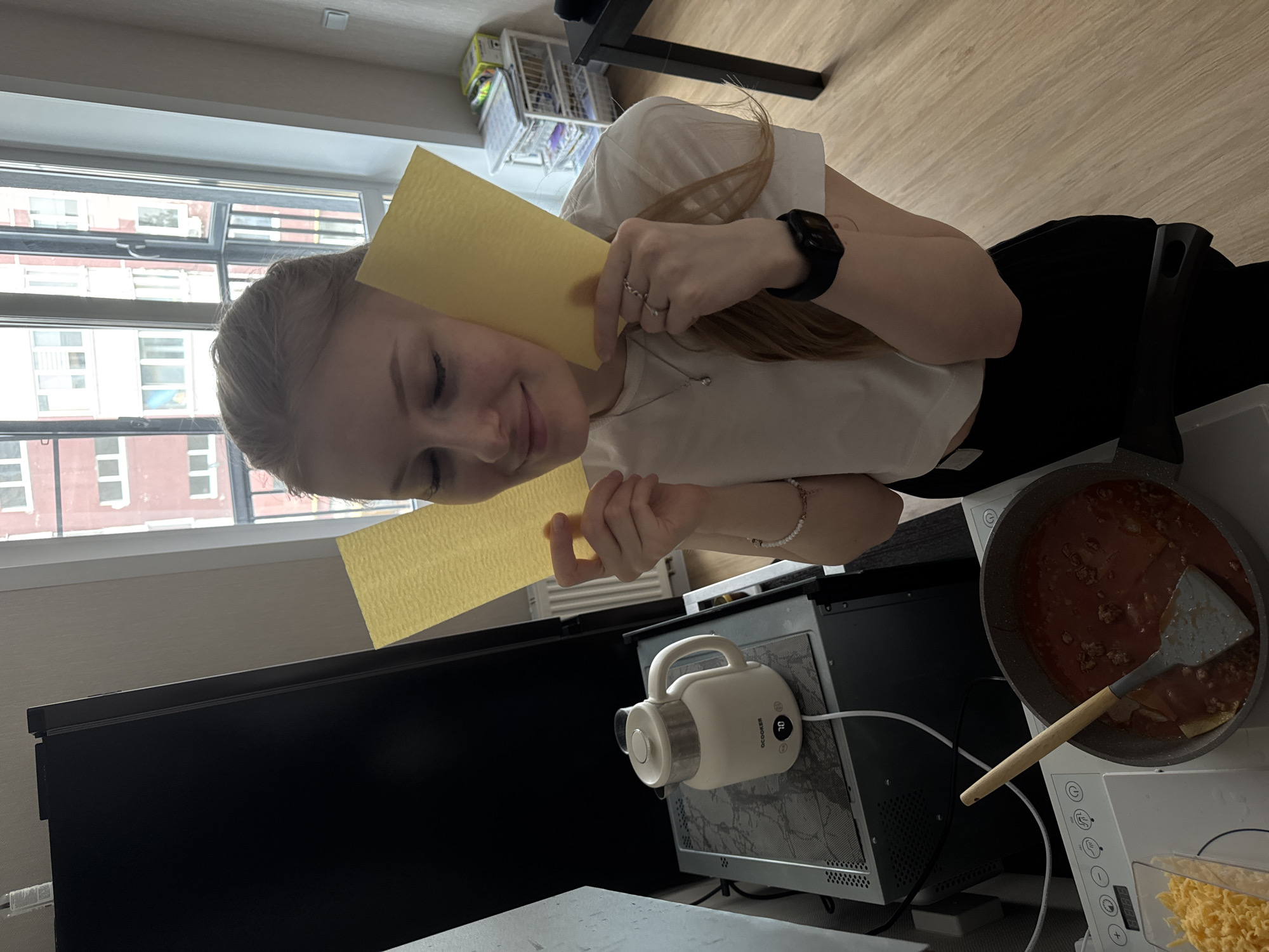 An indoor, sideways-oriented medium shot captures a young woman preparing food, likely lasagna, in what appears to be a home kitchen or living area in Perm, Russia. The woman, with light-colored hair, is seen from the chest up, smiling softly with her eyes closed. She wears a white t-shirt, black trousers, a black smartwatch on her left wrist, rings on her left hand, and a white beaded bracelet on her right. In her hands, she holds two rectangular, yellow lasagna noodles.
To her right, a dark grey frying pan contains a reddish-brown meat sauce, with a flat spatula resting within it. Below the pan, a clear container holds shredded yellow cheese. A white electric kettle, branded "OCOOKER" with "70" displayed on its digital screen, sits on a surface near a black monitor. In the background, a large window reveals multi-story urban buildings, indicating a daytime setting. A metal shelving unit is also partially visible. The overall scene conveys a sense of pleasant, domestic activity.