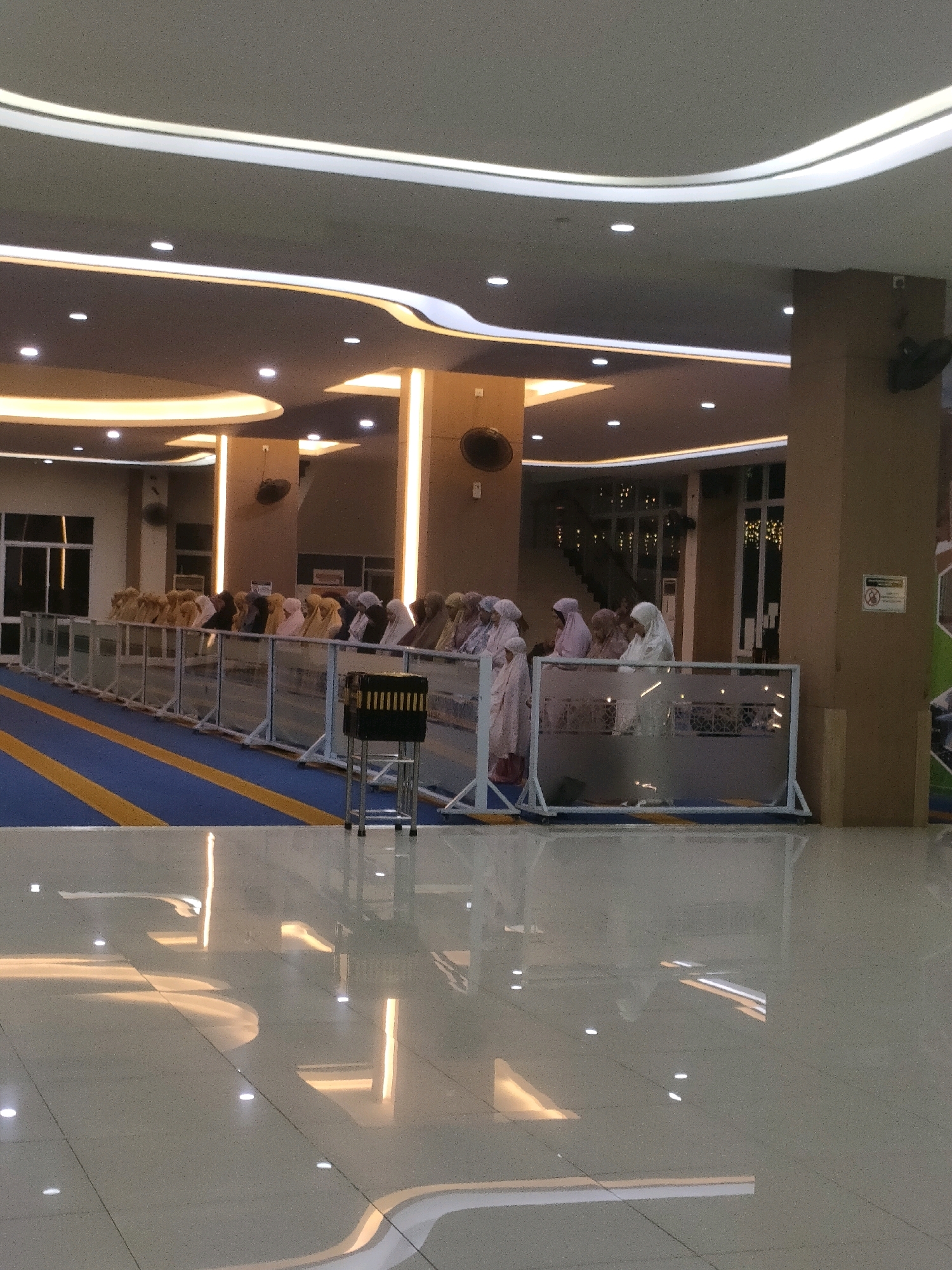 The image shows a group of people, predominantly women wearing hijabs and prayer clothes, engaged in prayer within a modern, indoor space. The setting appears to be a mosque or a prayer hall, characterized by its high ceilings, contemporary lighting fixtures, and reflective tiled floor. The prayer area is demarcated by a transparent railing, and a carpet with blue and yellow stripes is laid out in front of it. A small, black box with gold trim, possibly a donation box or a decorative item, stands in the foreground. The architectural style suggests a recent construction, with sleek lines and embedded lighting creating a warm ambiance. Based on the architectural style and typical indoor prayer settings in the region, this image is likely from a mosque in Batam, Indonesia. The lighting suggests it may be evening or early morning.