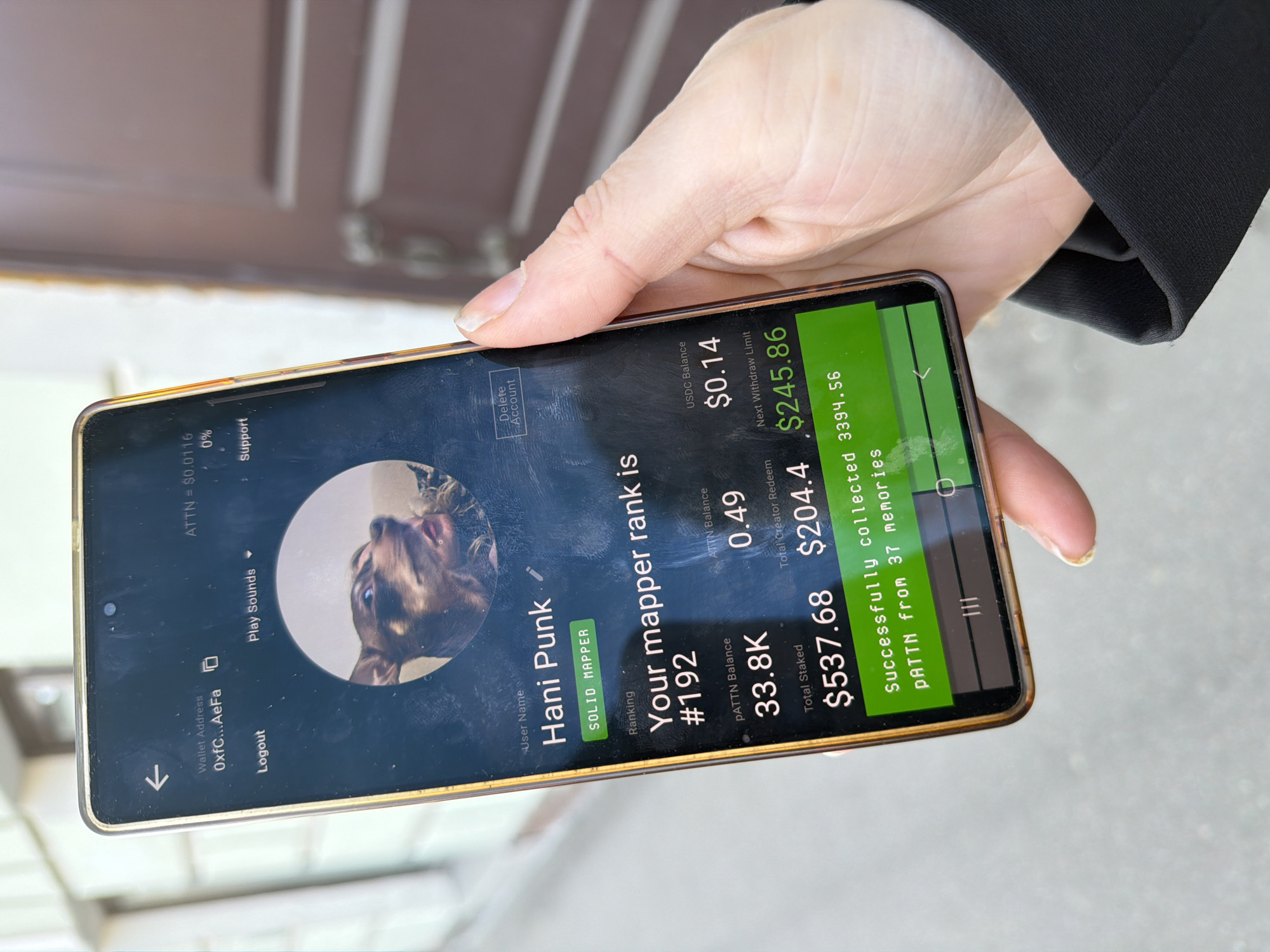 A person in Saint Petersburg, Russia, holds a smartphone displaying a screen showing the user profile "Hani Punk" on a cryptocurrency platform. The screen displays balances of various cryptocurrencies, including $0.14 in USDC, $0.49 in ATTN, and 33.8K in pATTN. The phone also indicates the user's mapper rank as #192. The total staked amount is $537.68 and the creator redeem is $204.4. The background shows a building exterior.