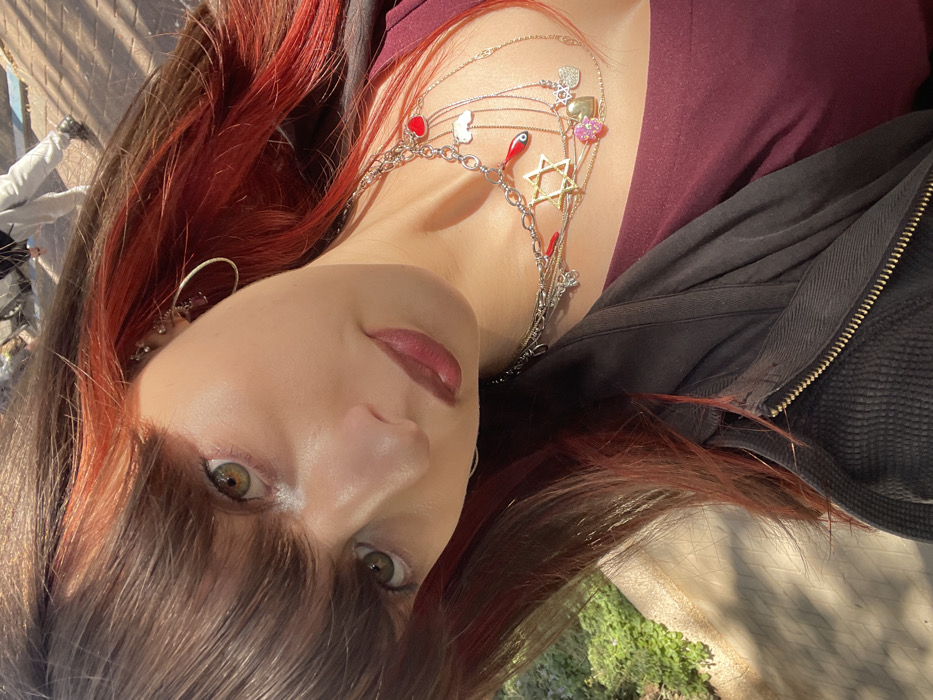 A close-up outdoor portrait displays a female subject with long, brown hair featuring prominent red streaks and bangs, looking directly into the camera. She has green/hazel eyes, a fair complexion, and wears a muted red lipstick. Her left ear is adorned with a small hoop earring and multiple stud piercings on the upper cartilage. She wears a layered necklace ensemble consisting of silver and gold-toned chains, bearing charms including a gold-colored Star of David, multiple red hearts, a blue evil eye, a silver Eiffel Tower, a pink floral cluster, and a silver heart. Her clothing comprises a maroon top and a dark, textured zippered jacket with a visible golden zipper on the right side. In the upper right background, the lower legs of an individual wearing light-colored trousers and dark shoes are partially visible on a sunlit paved surface with a blue line. The lower left background shows green foliage and a light-colored wall. The scene is set in the city of Raanana, Israel, under bright, sunny conditions.